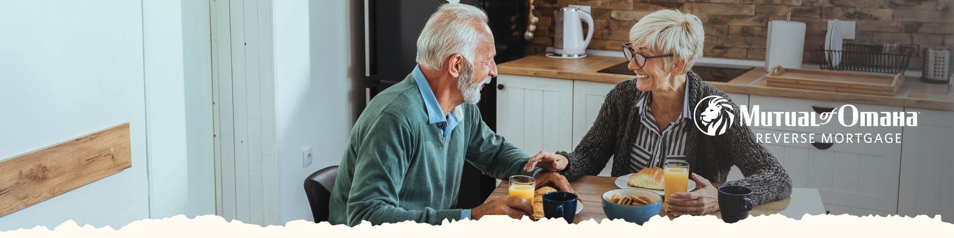 Two older adults talking at a table in a bright workshop, with tools and wood pieces nearby