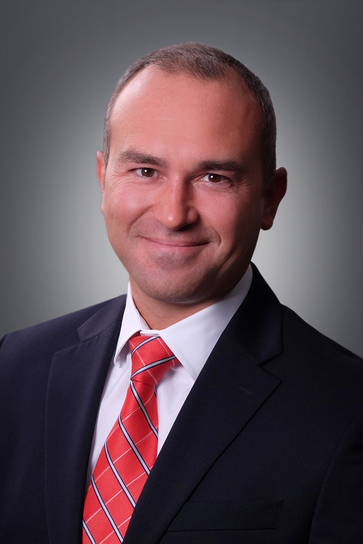 Formal portrait of a smiling man in a dark suit, white shirt, and red striped tie against a gray background