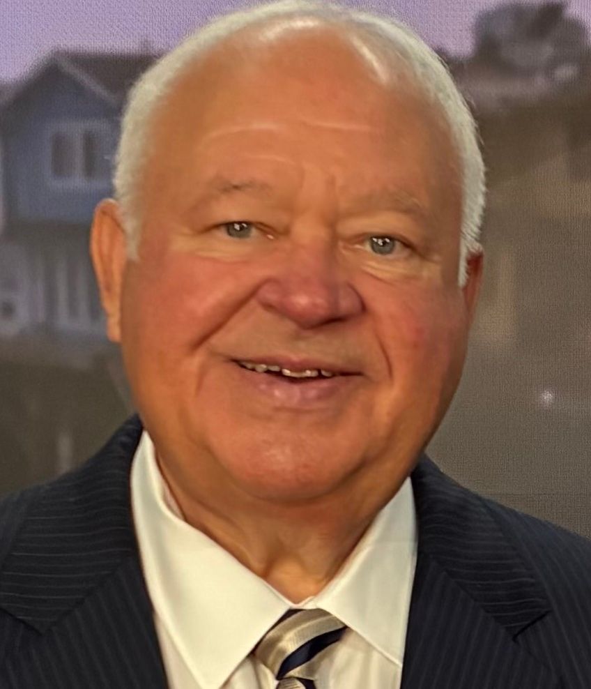 Formal portrait of a smiling man in a dark suit, white shirt, and red striped tie against a gray background