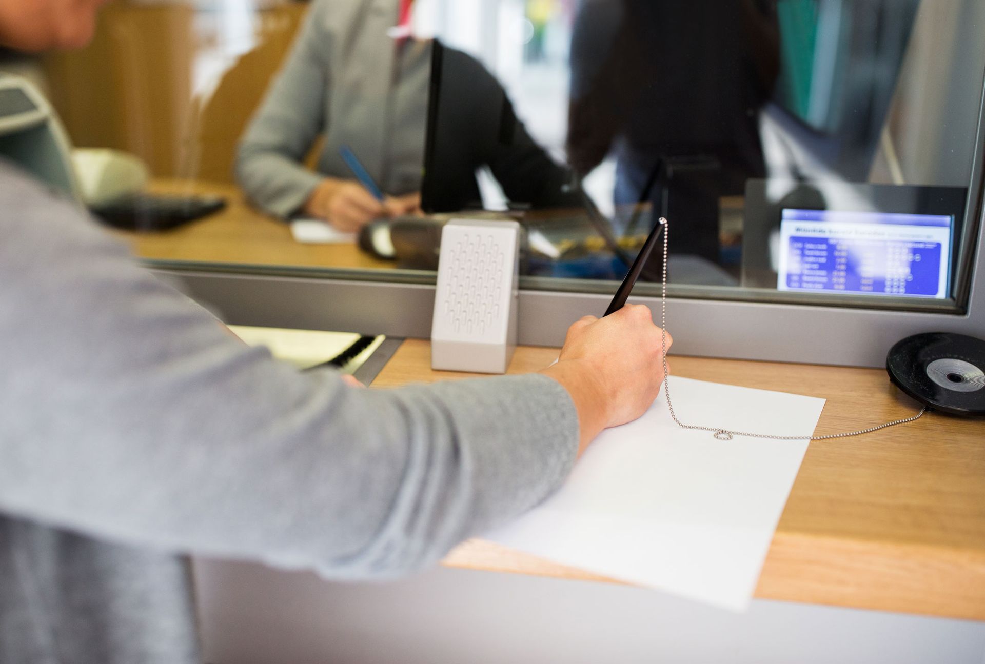 Person signing a document at a bank counter. Glass partition, bank employee partially visible.