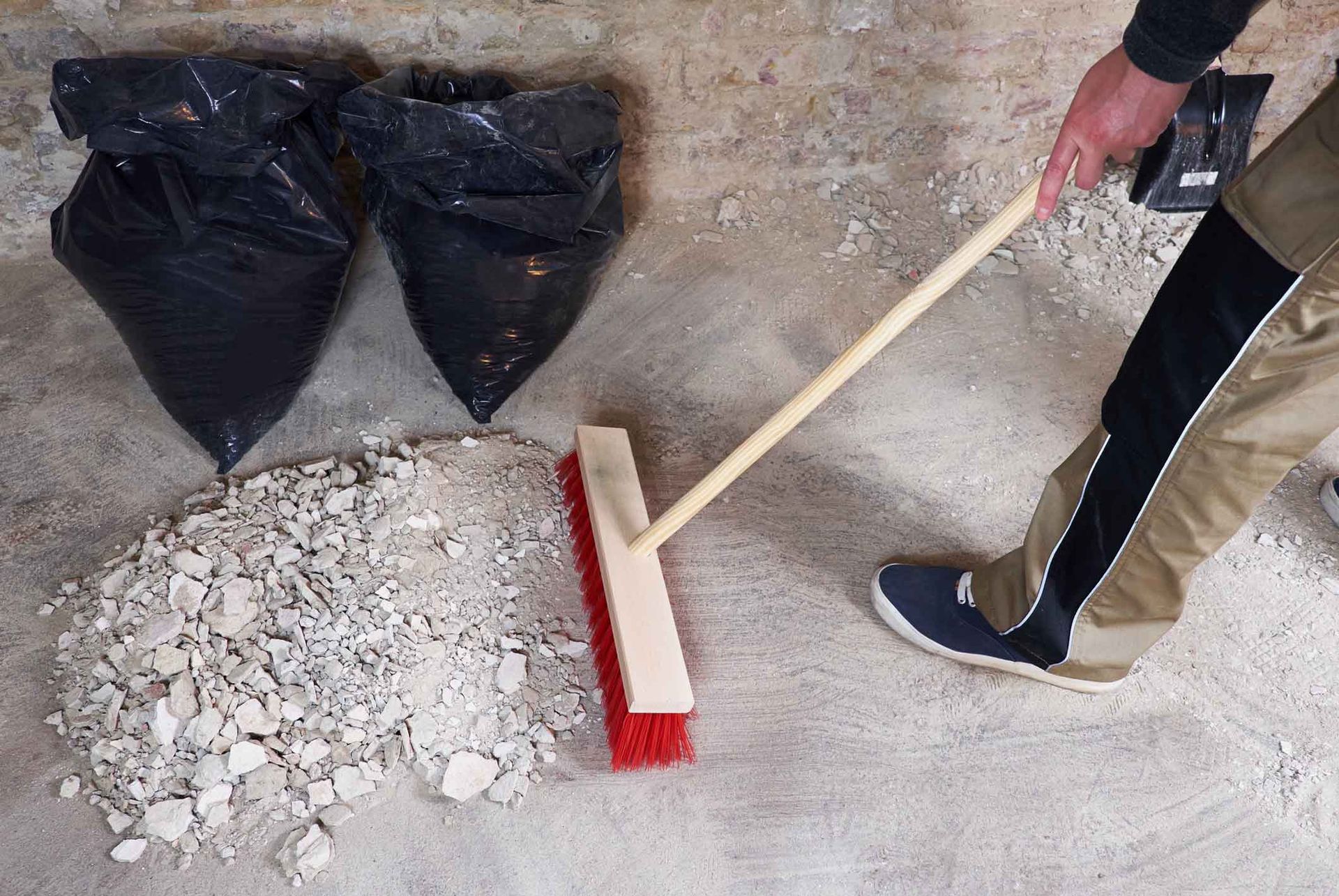 Person sweeping debris into a pile on a concrete floor with two trash bags nearby.
