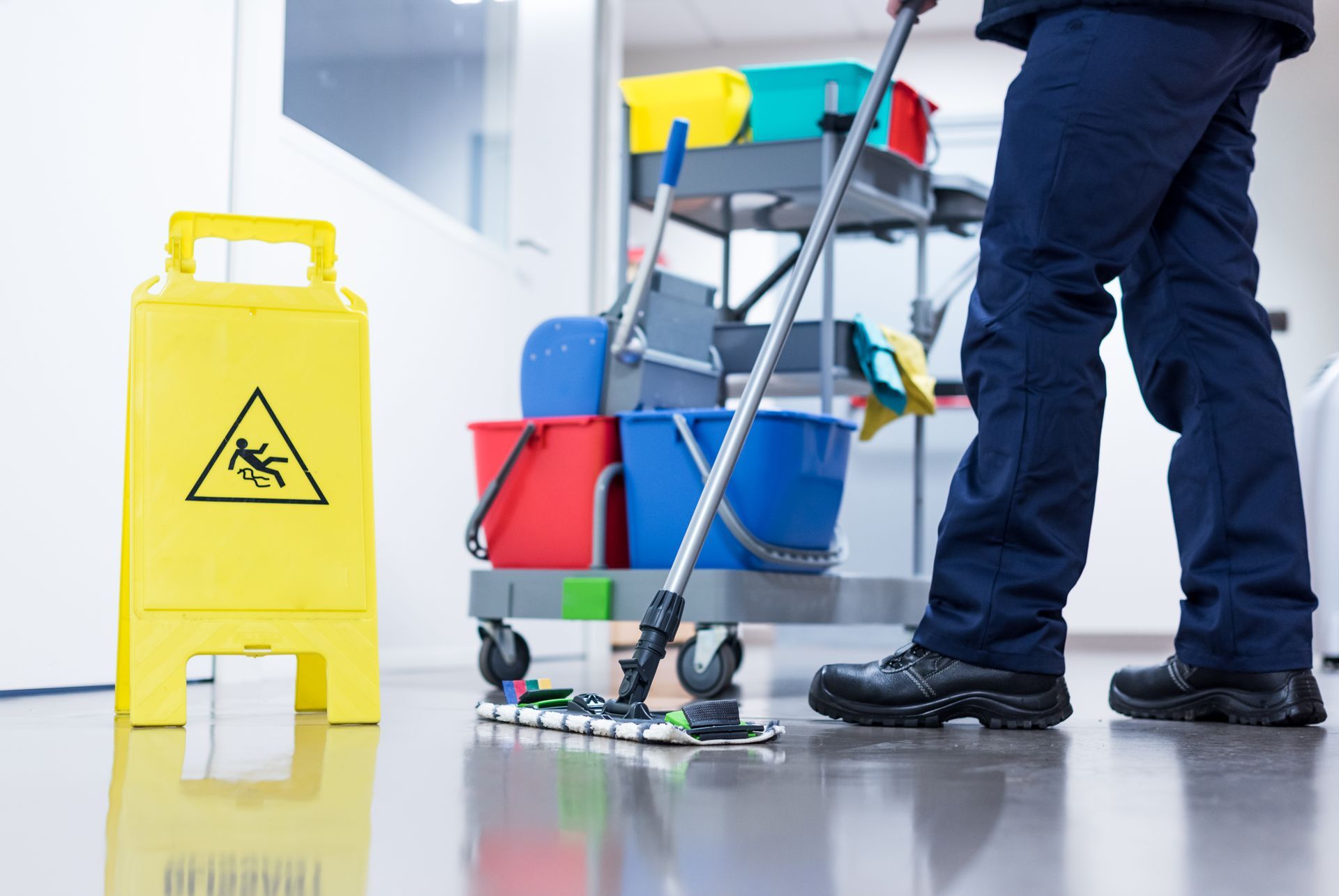 A person mopping a wet floor next to a yellow caution sign and cleaning cart. A person mopping a wet floor next to a yellow caution sign and cleaning cart.