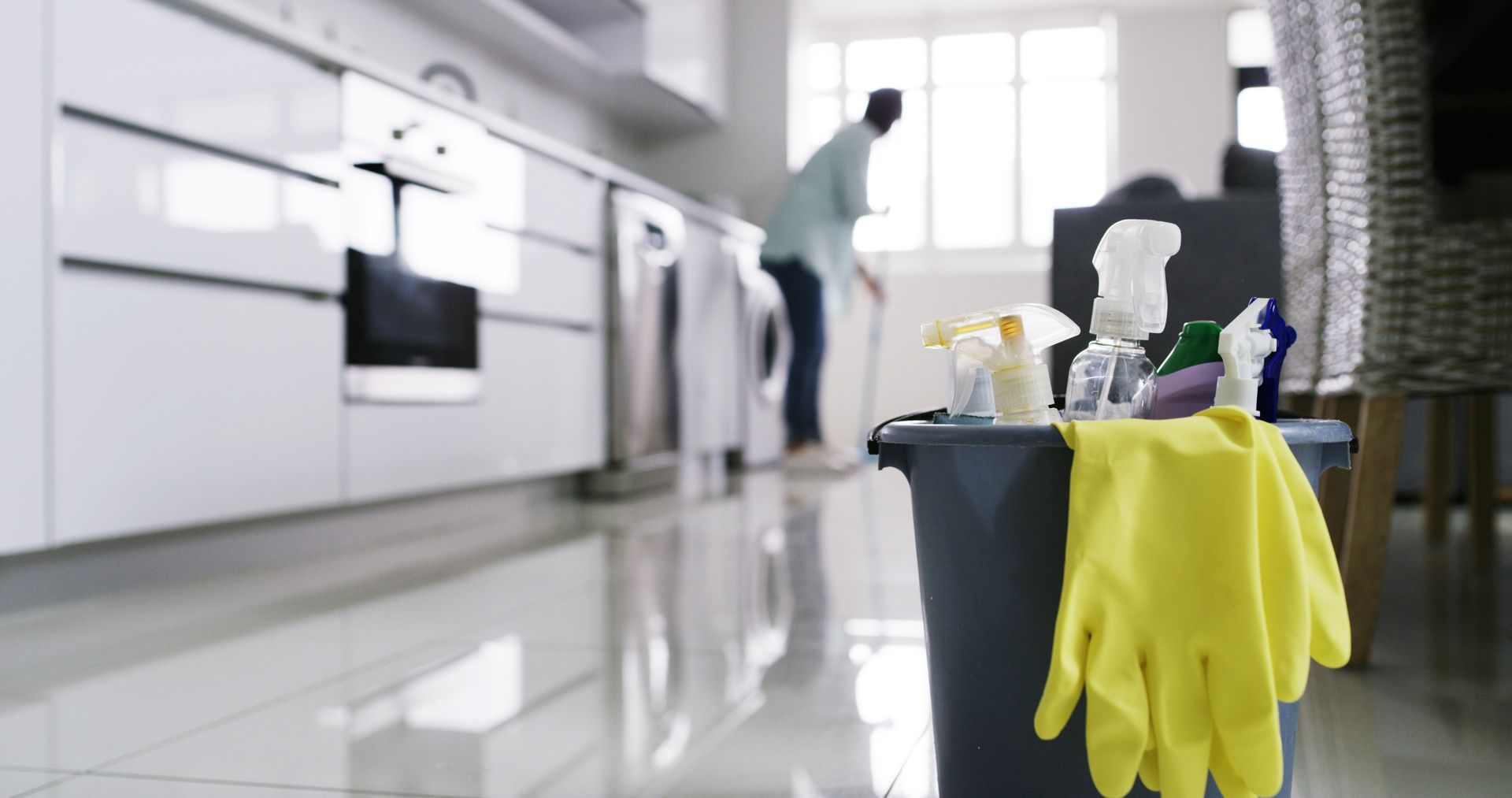 Cleaning supplies with yellow gloves on a bucket in a bright kitchen while a person cleans in the background Cleaning supplies with yellow gloves on a bucket in a bright kitchen while a person cleans in the background