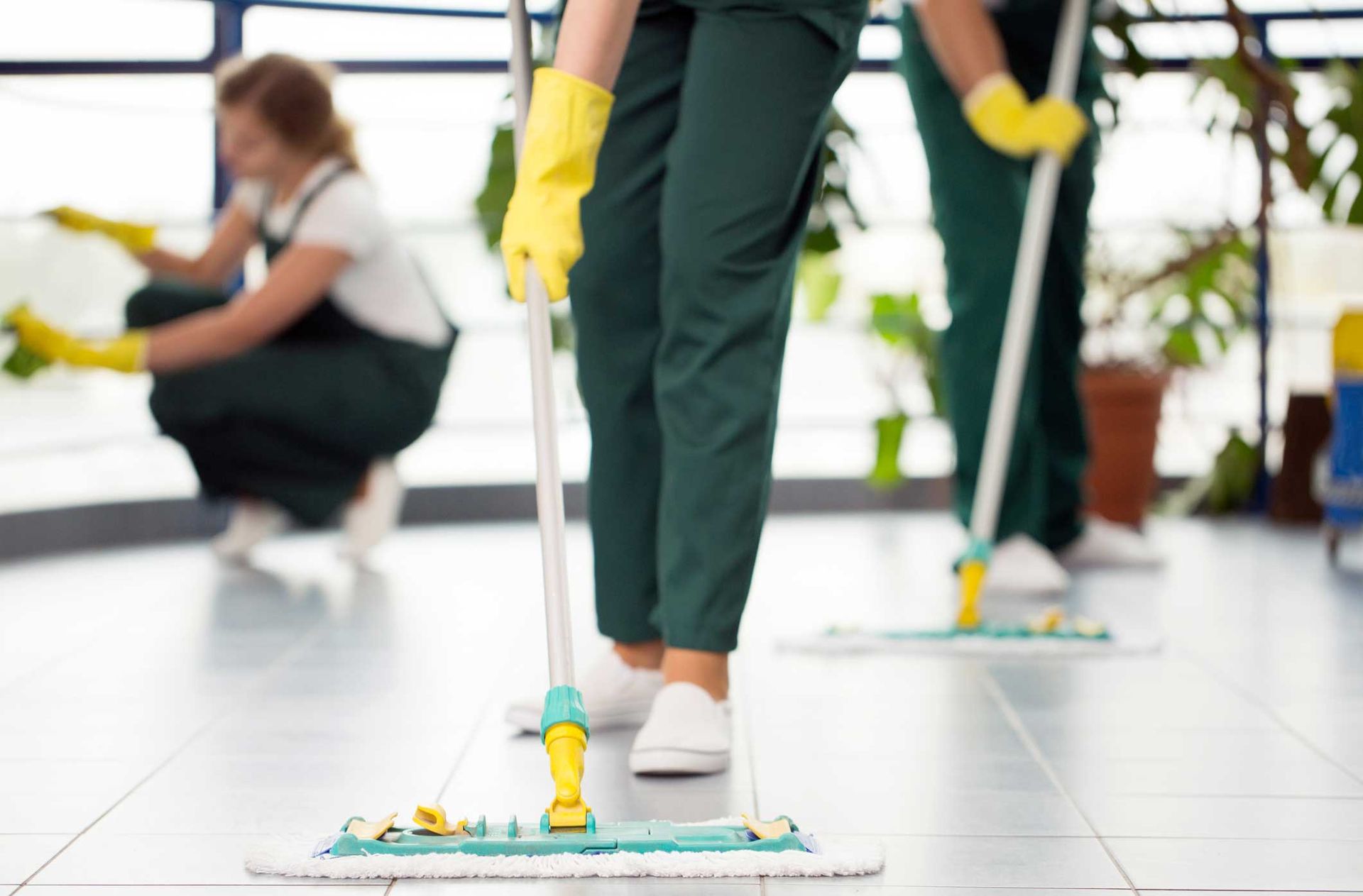 Janitors mopping a tiled floor in a bright indoor space, with yellow gloves and cleaning tools