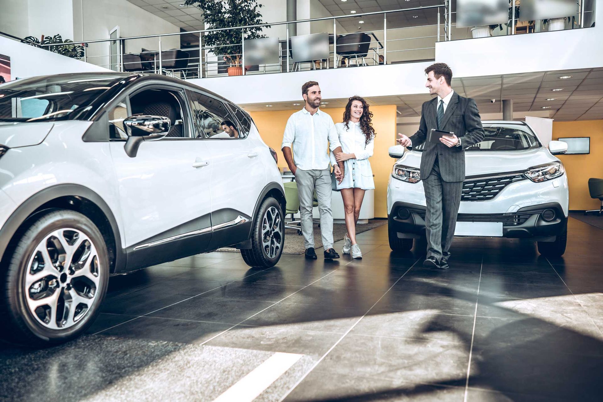 Three people stand beside two white SUVs in a bright car showroom.