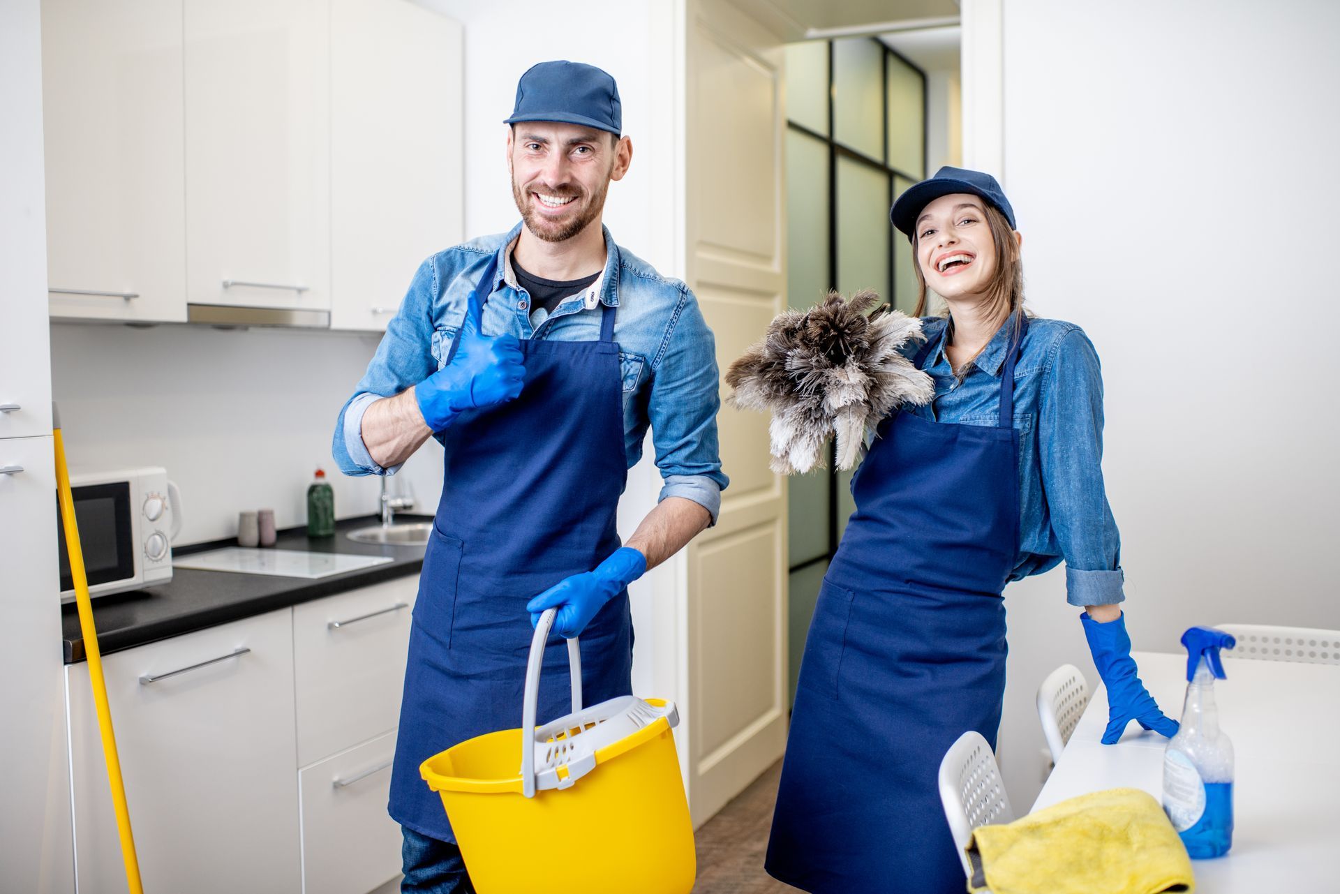 Two cleaners in blue uniforms holding cleaning supplies and a mop in a bright kitchen Two cleaners in blue uniforms holding cleaning supplies and a mop in a bright kitchen