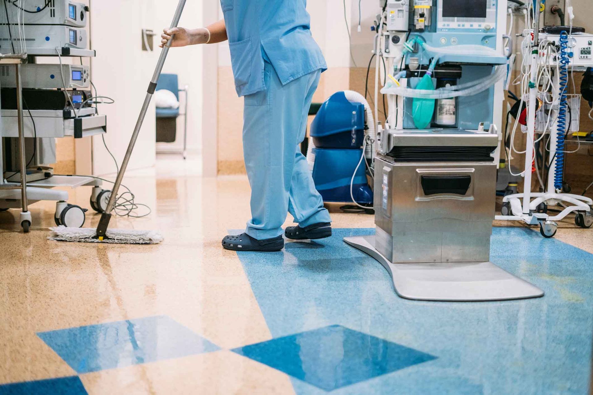 Hospital worker mopping a clinical floor beside medical equipment and a cart