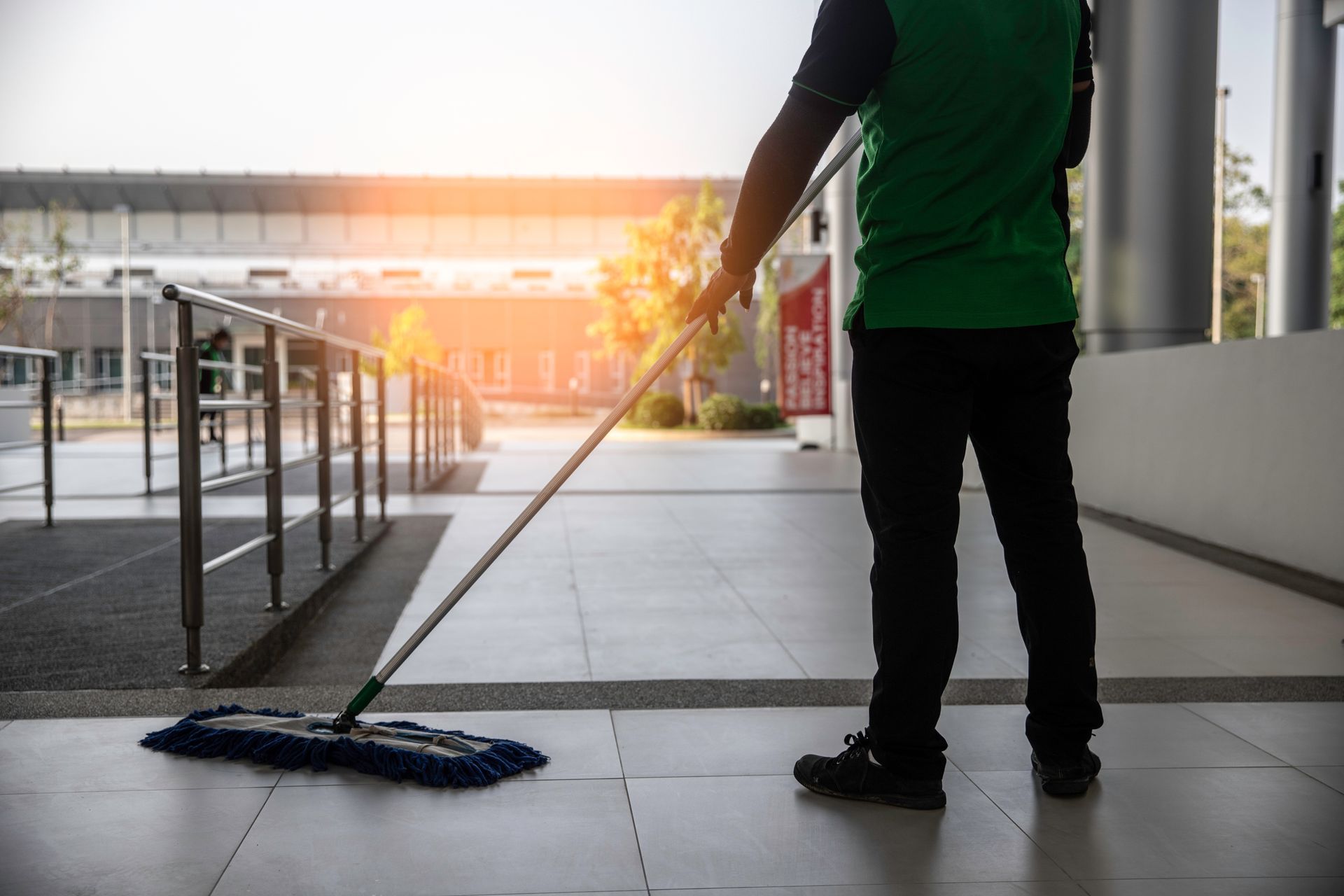 Person mopping a tile floor in a building, wearing a green shirt and black pants.