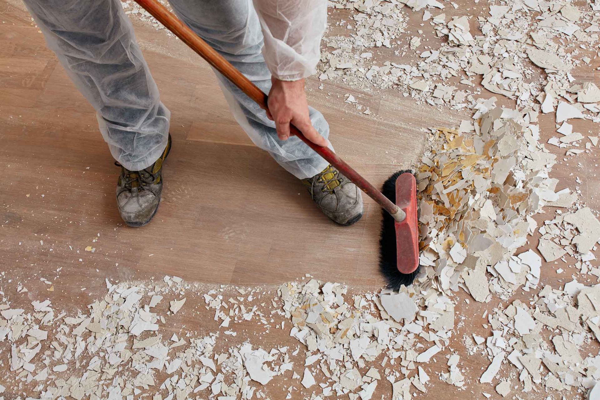 Person sweeping peeling paint off a wooden floor with a broom and dustpan