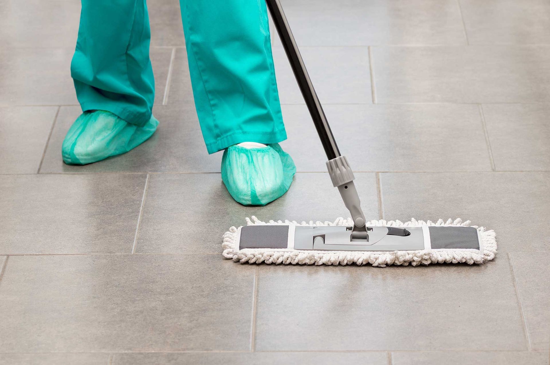 A person in teal shoes mops a gray tiled floor with a flat mop.