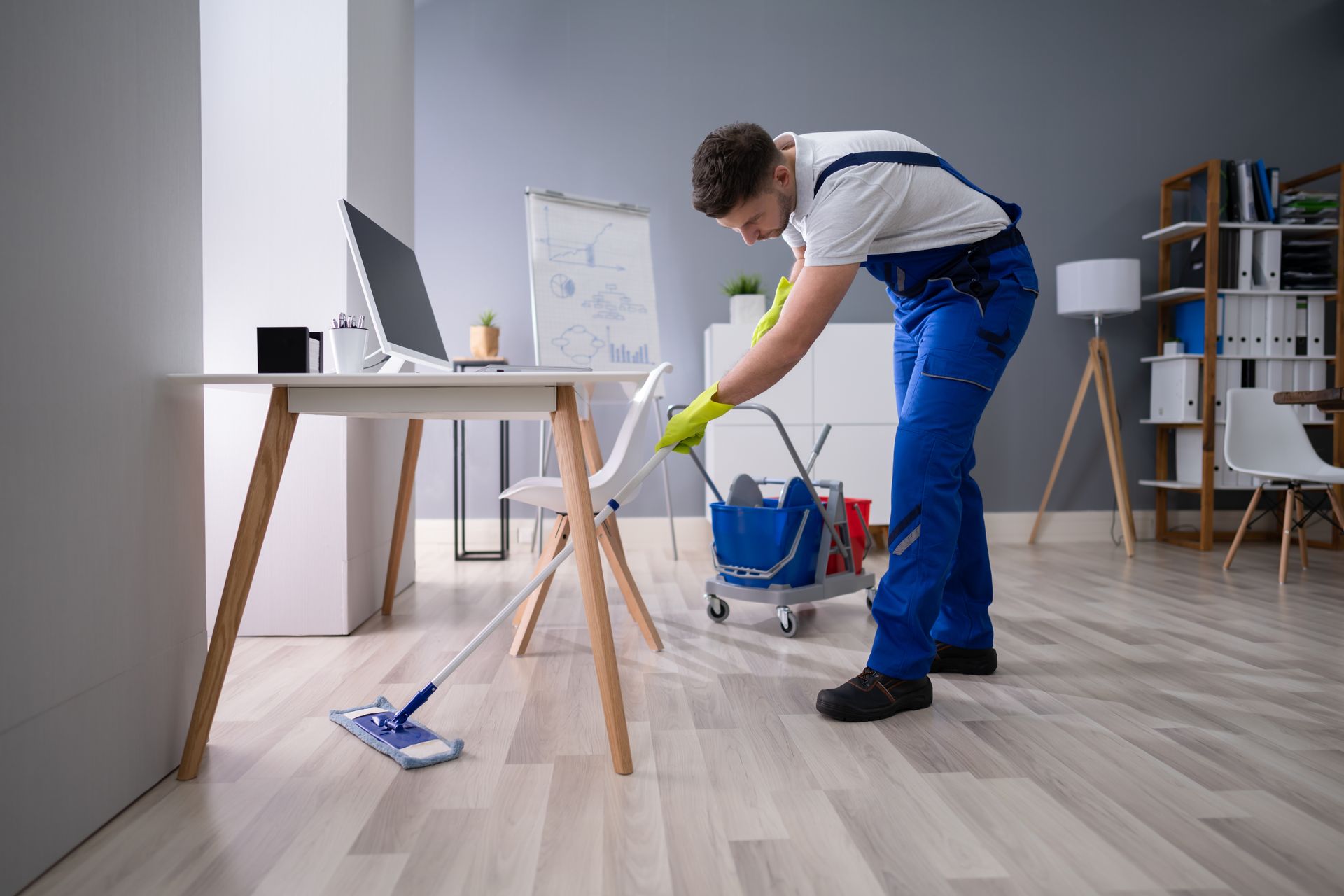 Person mopping a light wood floor in a bright room with a blue bucket and cleaning supplies Person mopping a light wood floor in a bright room with a blue bucket and cleaning supplies