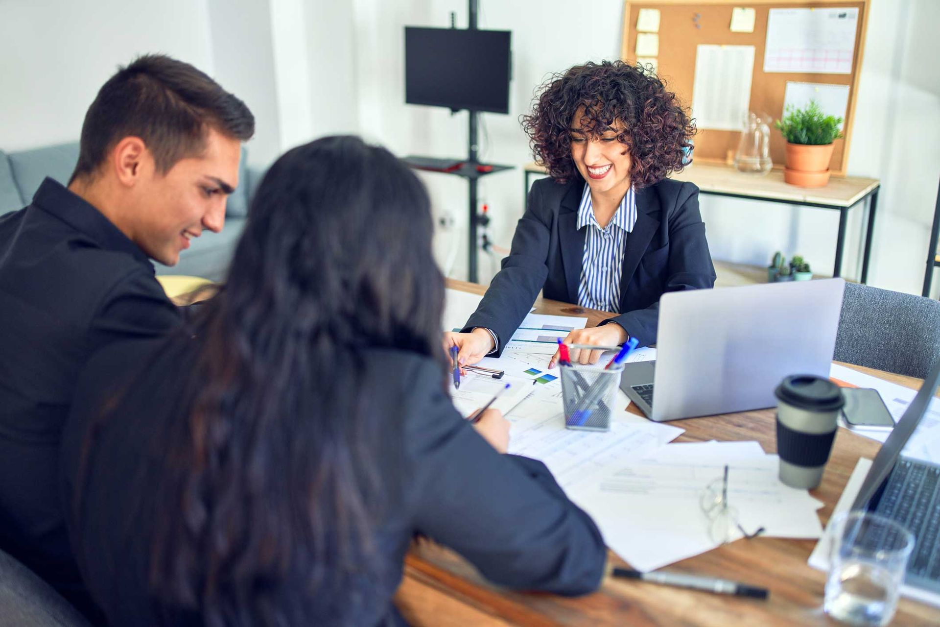 Three people in business attire at a table, smiling and reviewing documents in an office.