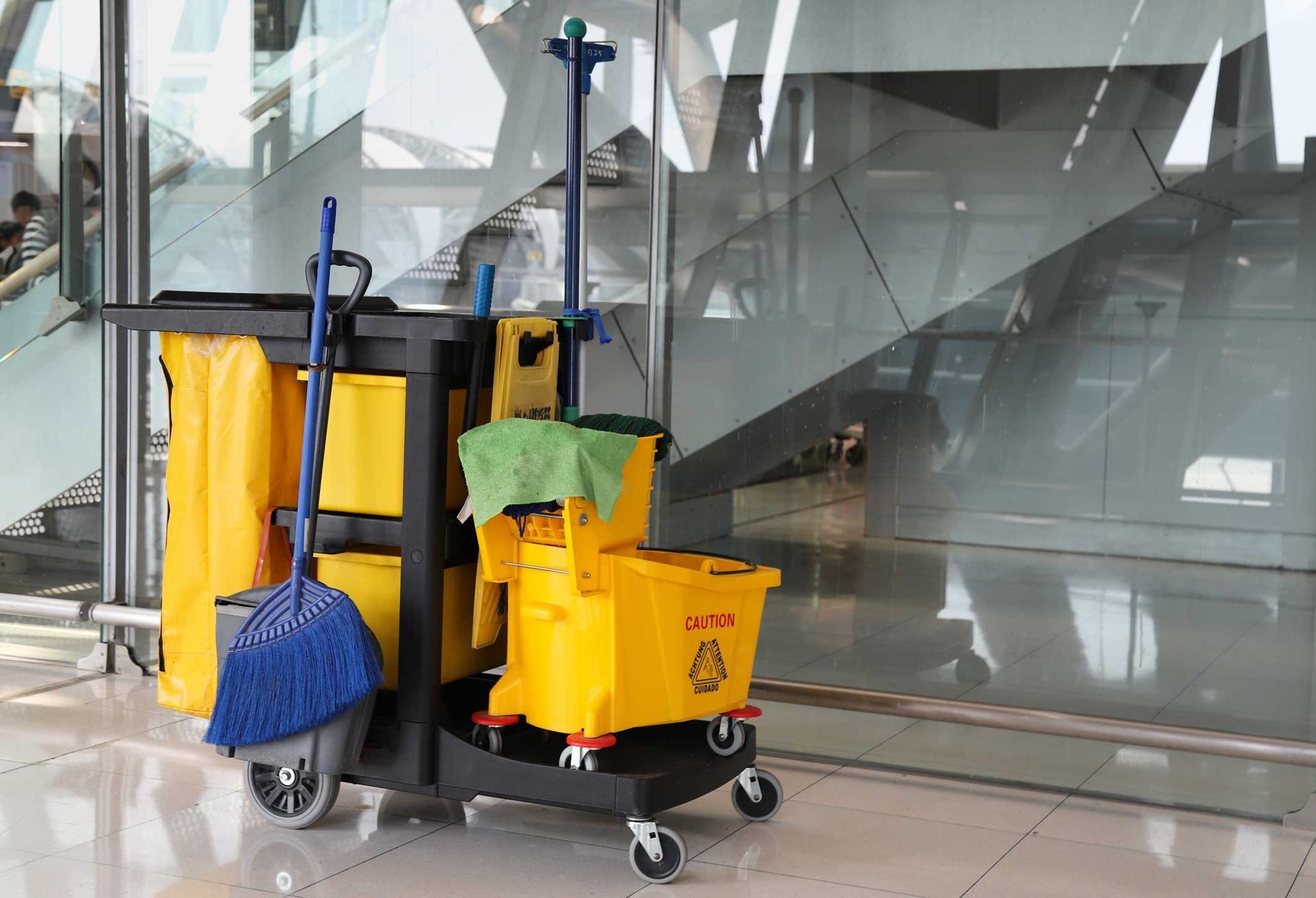 Yellow cleaning cart with buckets, mop, and broom on wheels, near a glass wall.