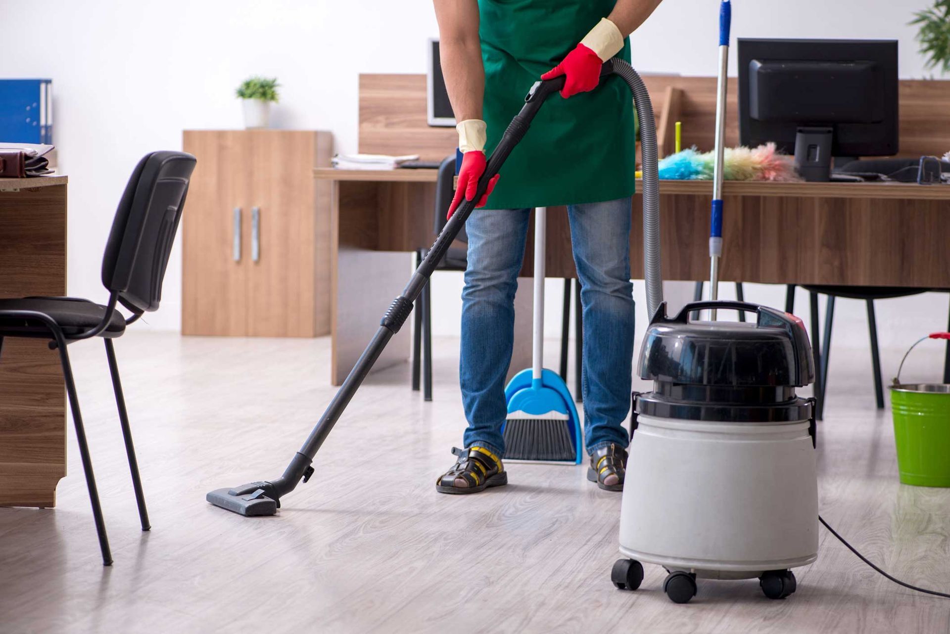 Person vacuuming an office floor, wearing an apron and gloves. A vacuum cleaner and cleaning supplies are nearby. Person vacuuming an office floor, wearing an apron and gloves. A vacuum cleaner and cleaning supplies are nearby.