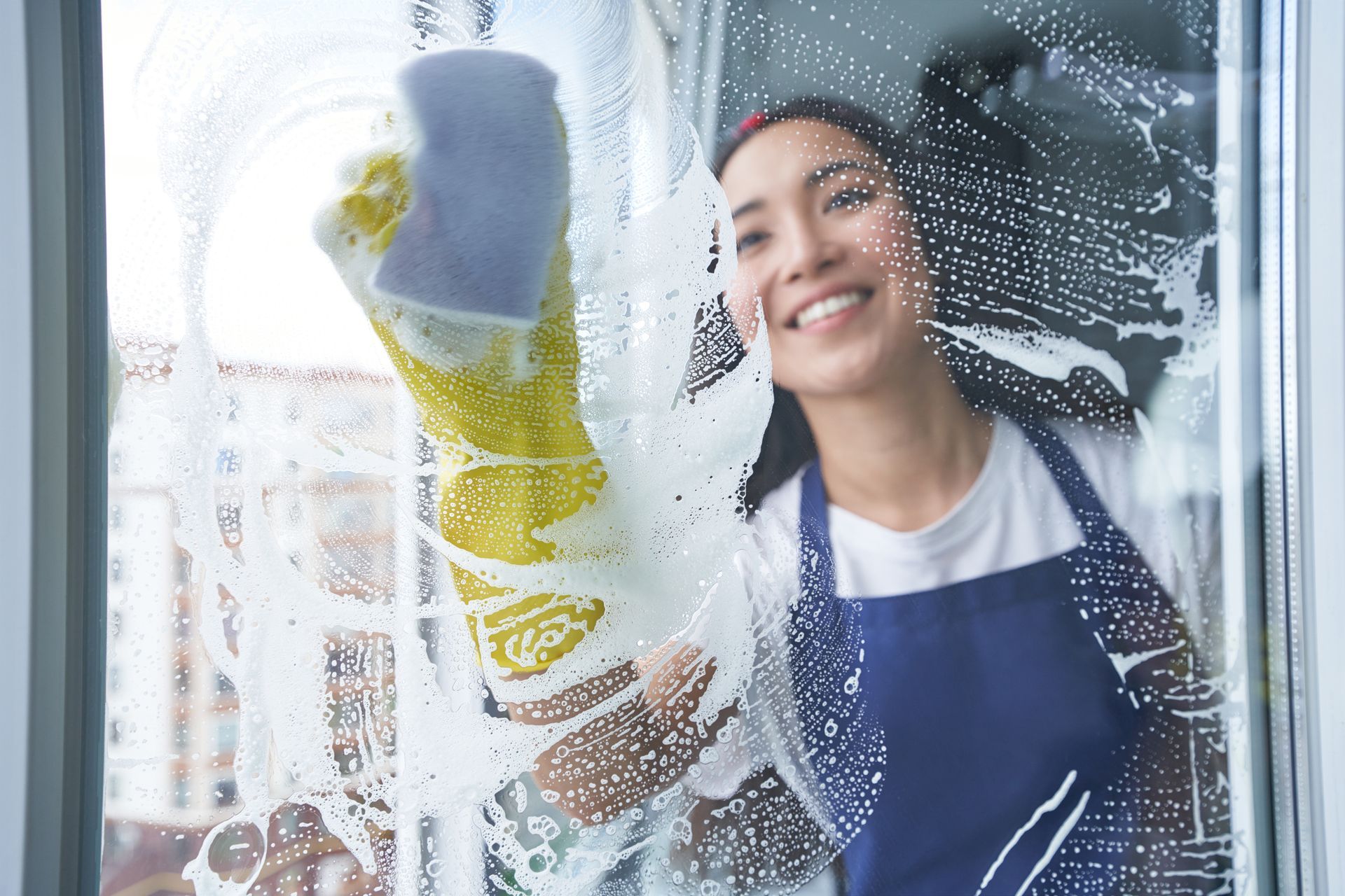 Woman cleaning a window with a sponge, smiling, with soap suds and water streaks on the glass.