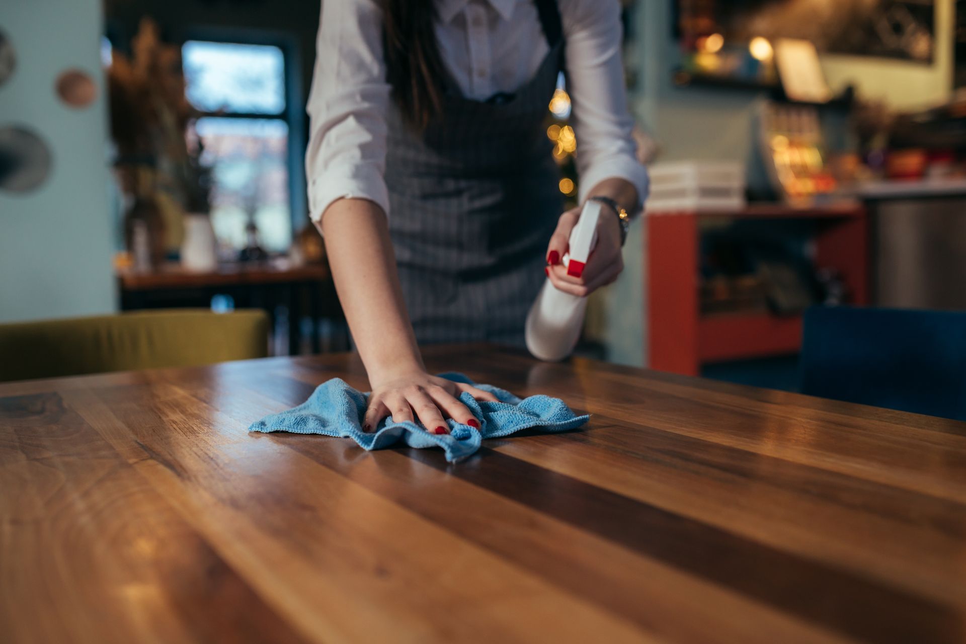 Person wiping a wooden table with a blue cloth and spray bottle in a restaurant. Person wiping a wooden table with a blue cloth and spray bottle in a restaurant.