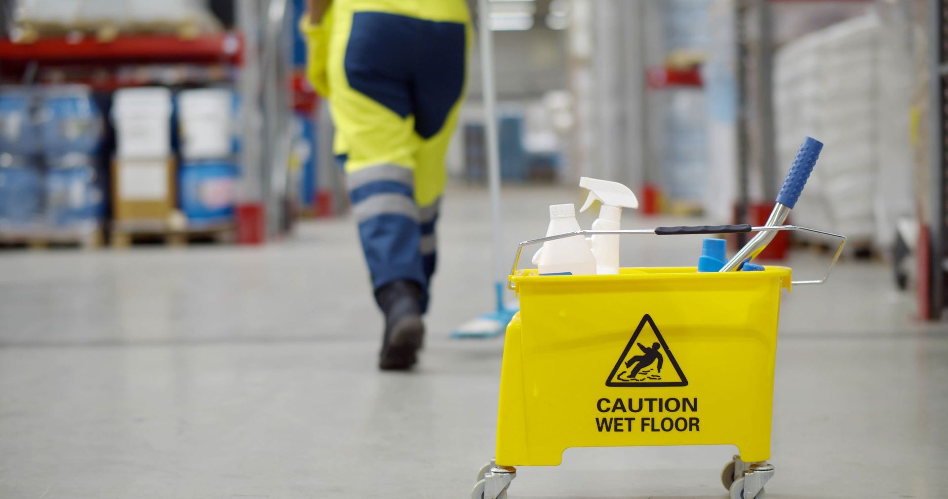 Yellow wet floor caution sign with cleaning supplies in a warehouse aisle, worker walking away in background
