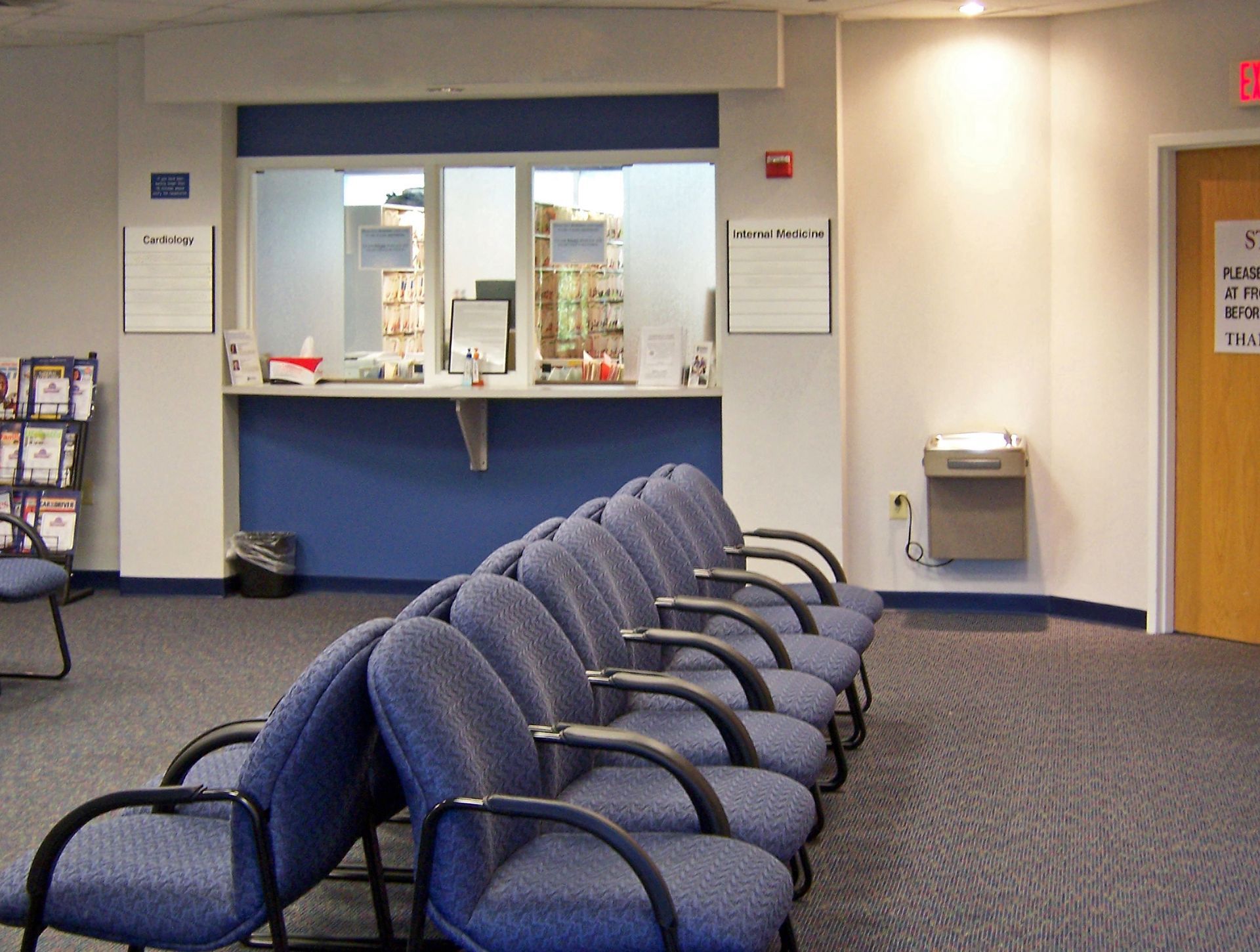A row of blue chairs faces a service counter in a quiet office waiting area with a water fountain and a wooden door.