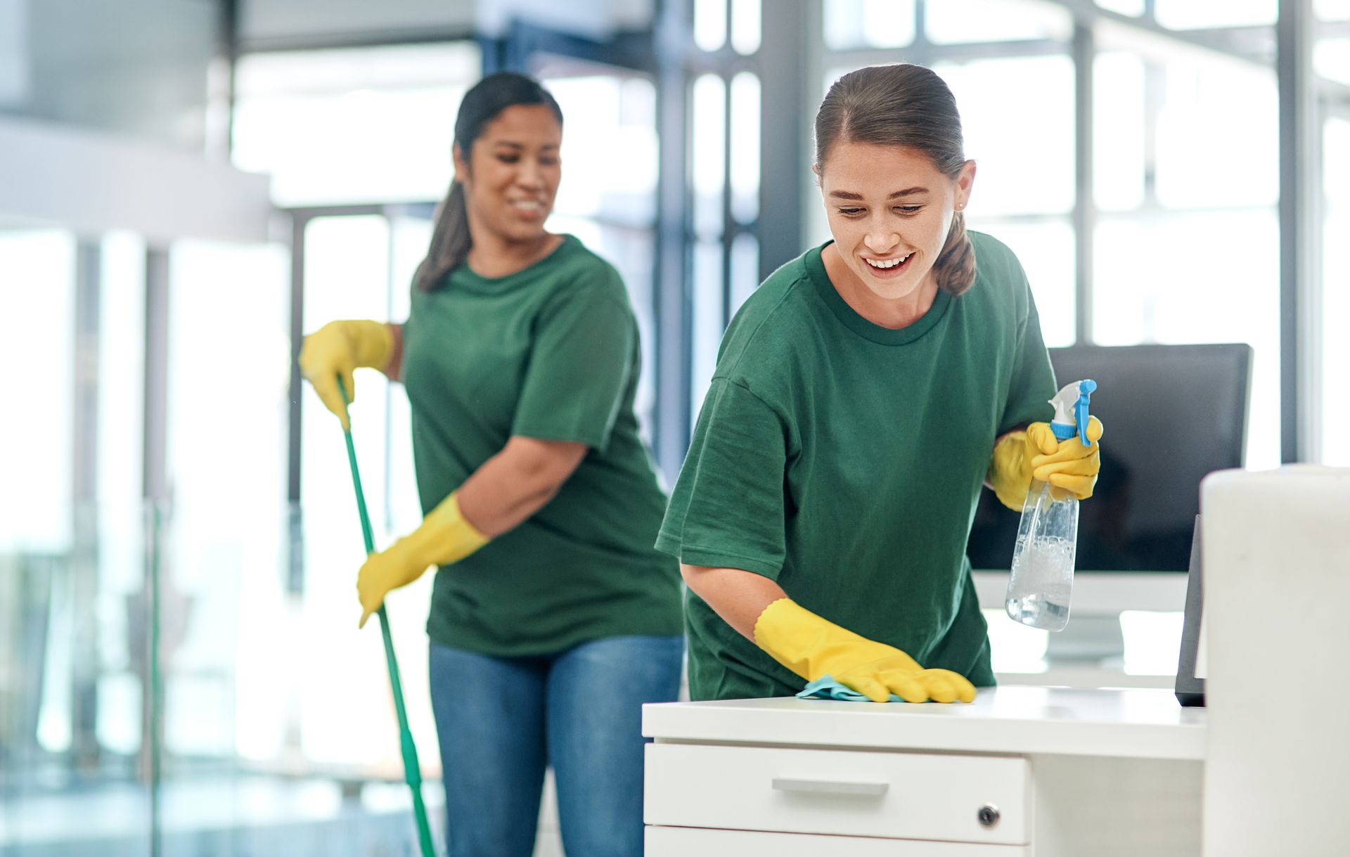 Two people cleaning an office: One mopping, one wiping a desk, both in yellow gloves and green shirts. Two people cleaning an office: One mopping, one wiping a desk, both in yellow gloves and green shirts.