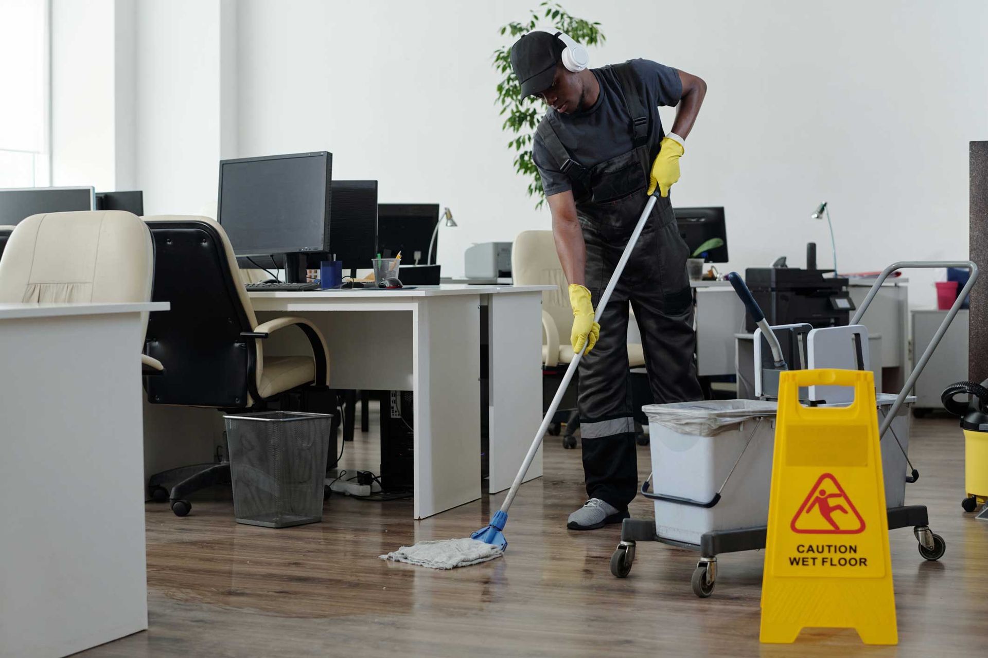 Cleaner mopping office floor beside desks and a yellow wet floor sign