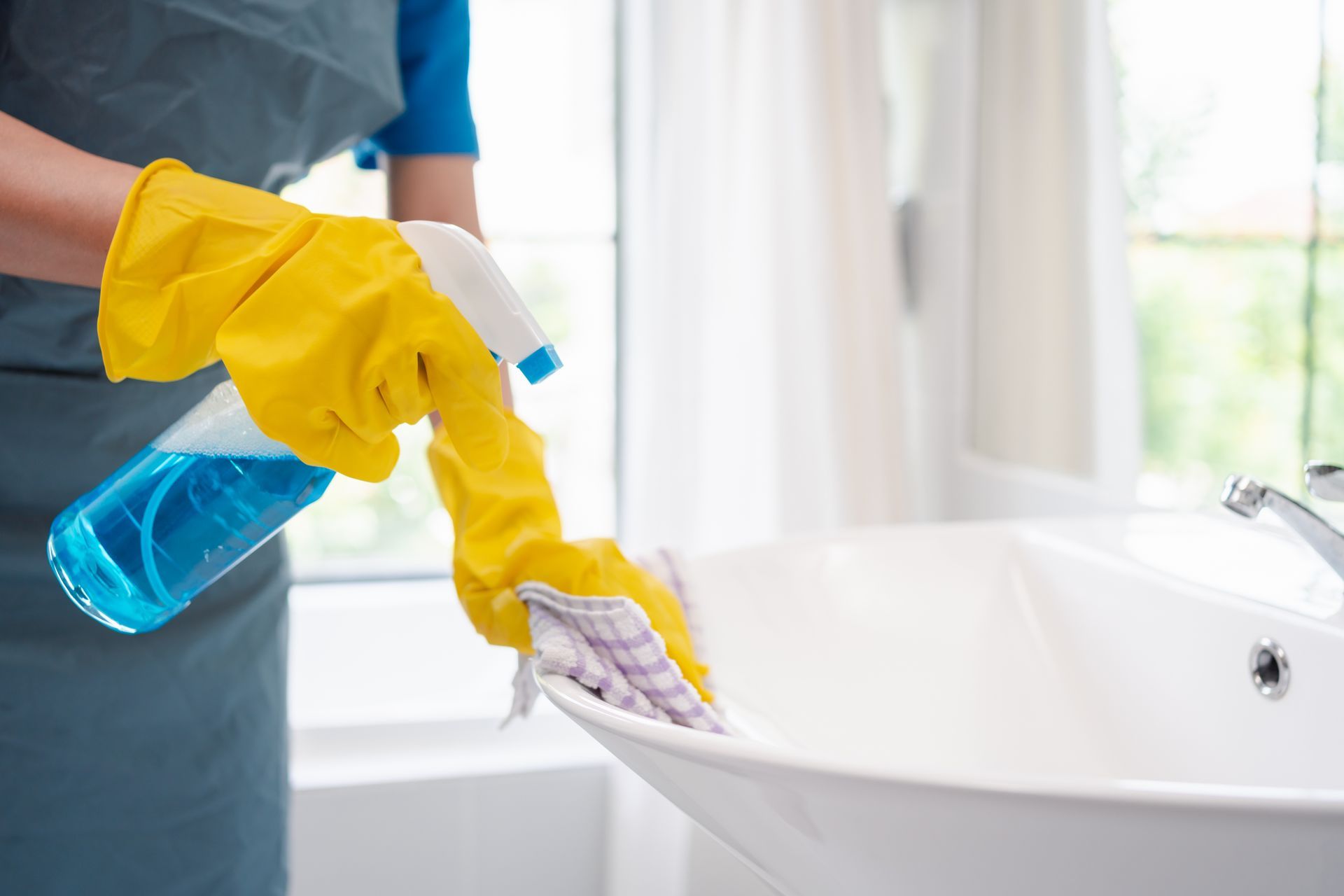 Person in yellow gloves spraying and wiping a white bathroom sink with a spray bottle and cloth.
