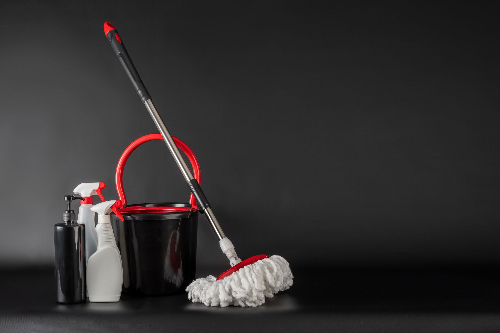 Cleaning supplies in a black bucket: mop, spray bottle, brush, and cloth on a dark background