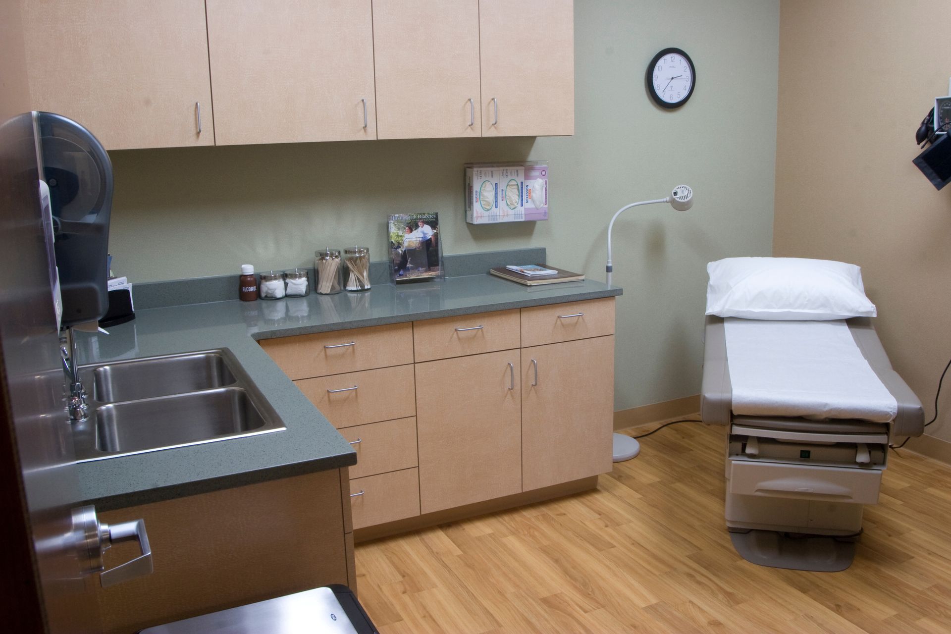 A clean medical exam room featuring a paper-covered exam table, a sink with light wood cabinets, and a wall-mounted clock.