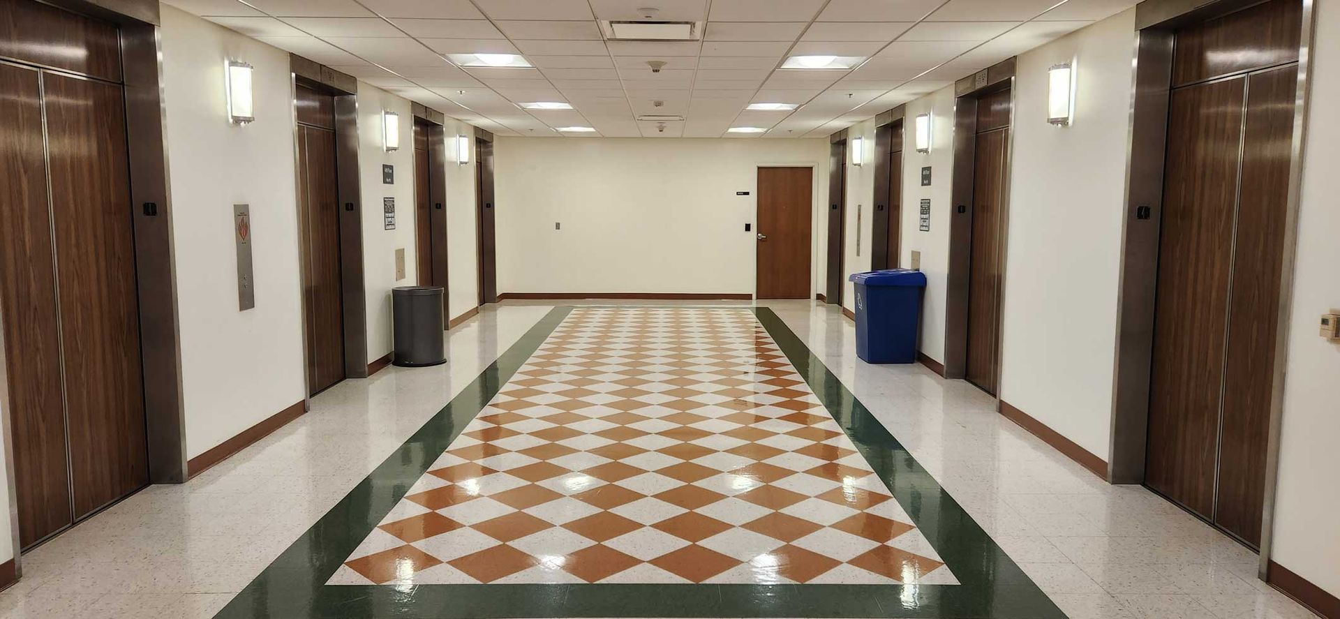 Empty hotel hallway with patterned tile floor, beige walls, wooden doors, and blue trash bins Empty hotel hallway with patterned tile floor, beige walls, wooden doors, and blue trash bins