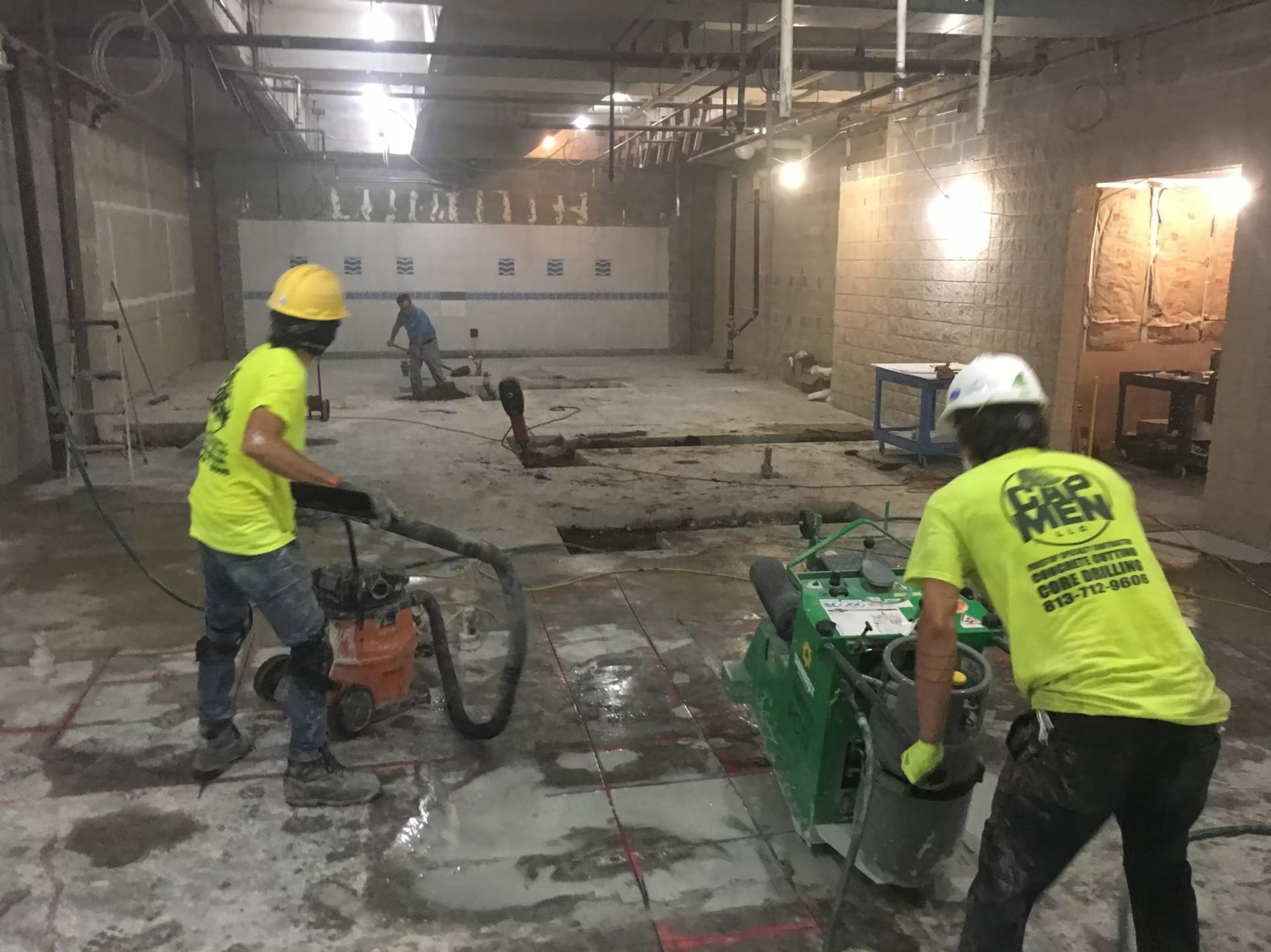 Workers operating construction equipment in a large indoor concrete space with bright safety shirts and hard hats