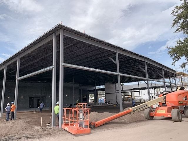Steel building under construction with workers and an orange boom lift outside.