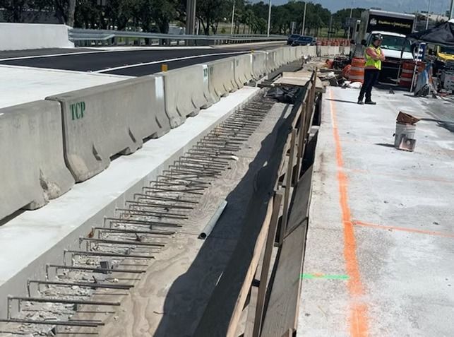 Road construction site with concrete barriers, exposed rebar, and workers near paving equipment
