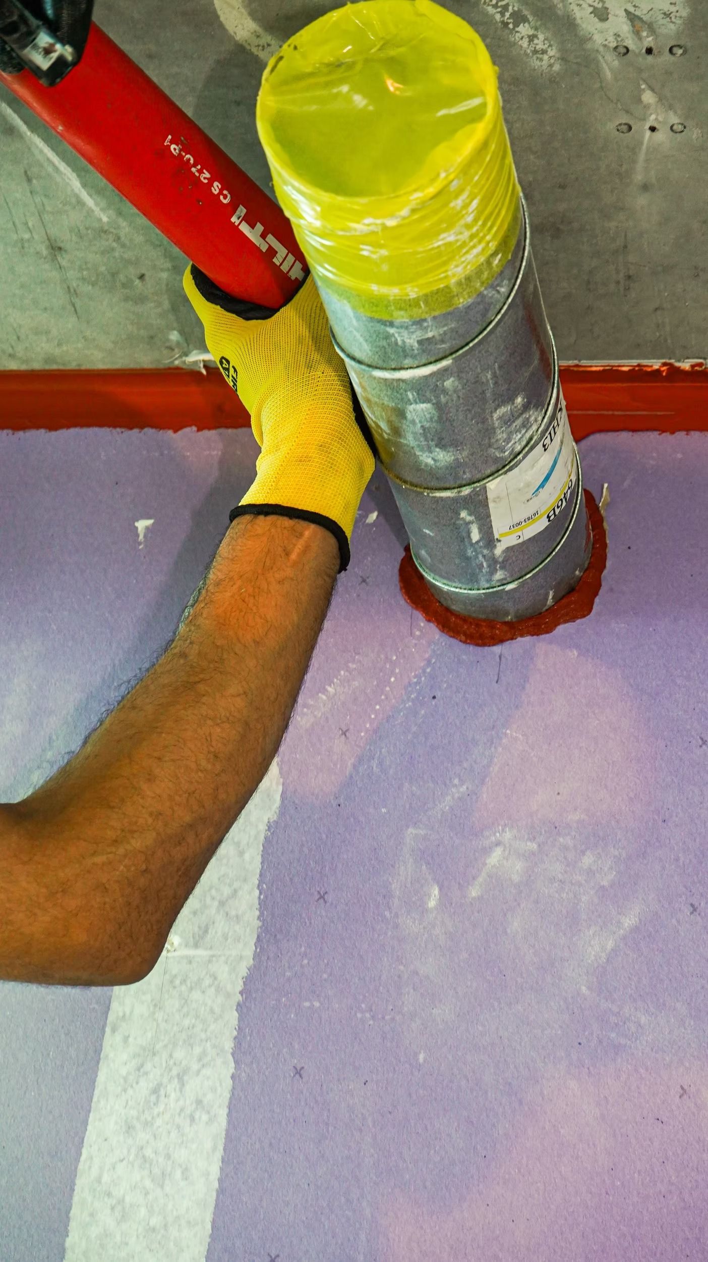 Gloved hand holding a wrapped pipe joint with red pipes against a purple floor and gray wall