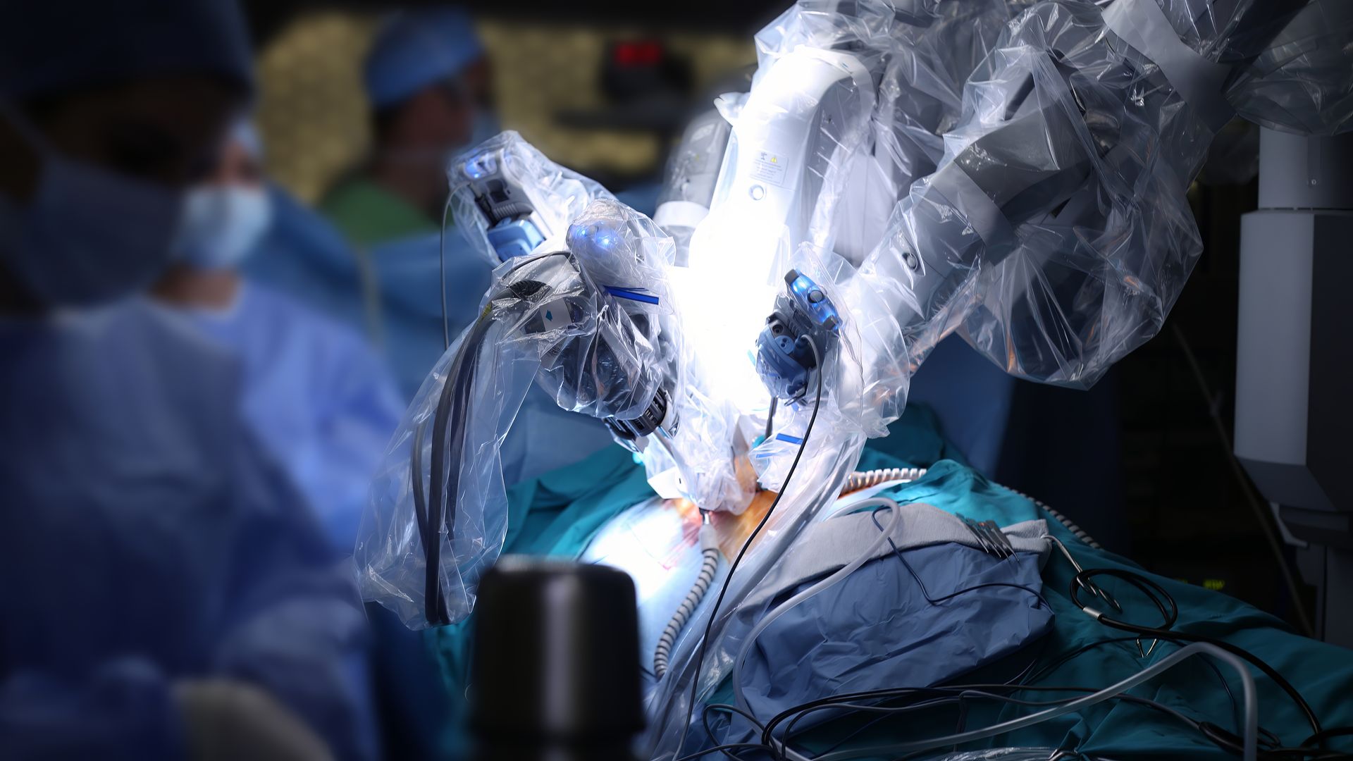 Cirurgia robótica em andamento, com cirurgiões de jaleco azul operando ao lado de um paciente coberto com um pano verde-água.