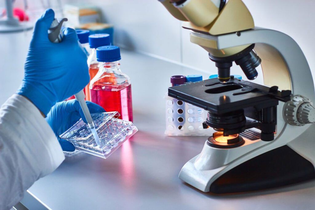Técnico de laboratório com luvas utilizando um microscópio ao lado de tubos de ensaio e uma pipeta e