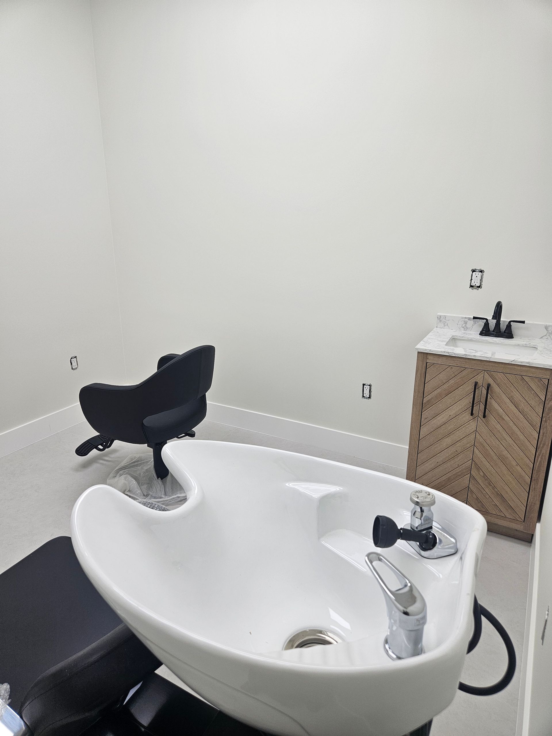 Modern salon shampoo bowl with black chair in a white room, beside a wicker cabinet and sink.