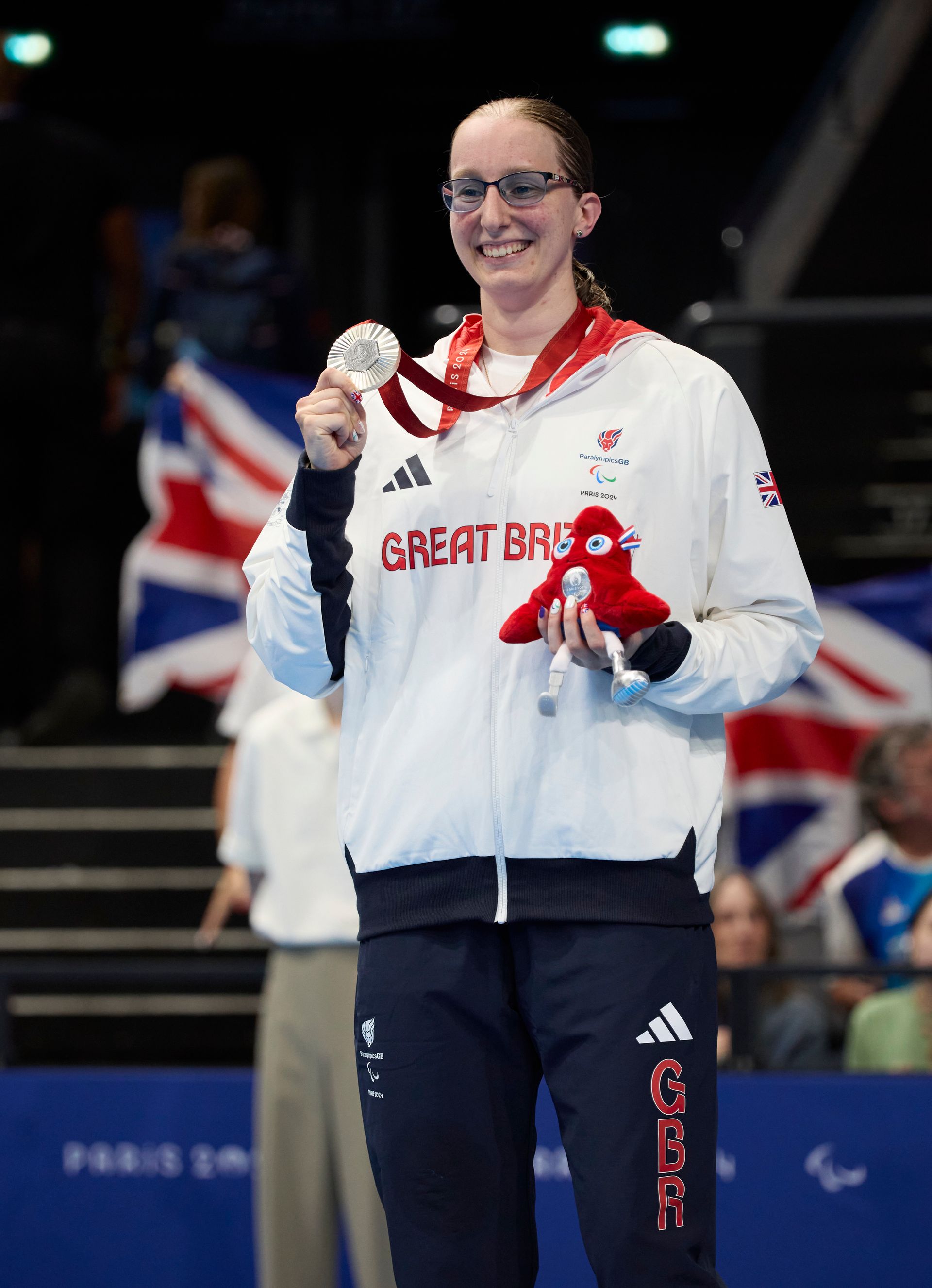 callie-ann has brown hair and wears glasses. she has a white jacket on with Great Britain in red wri
