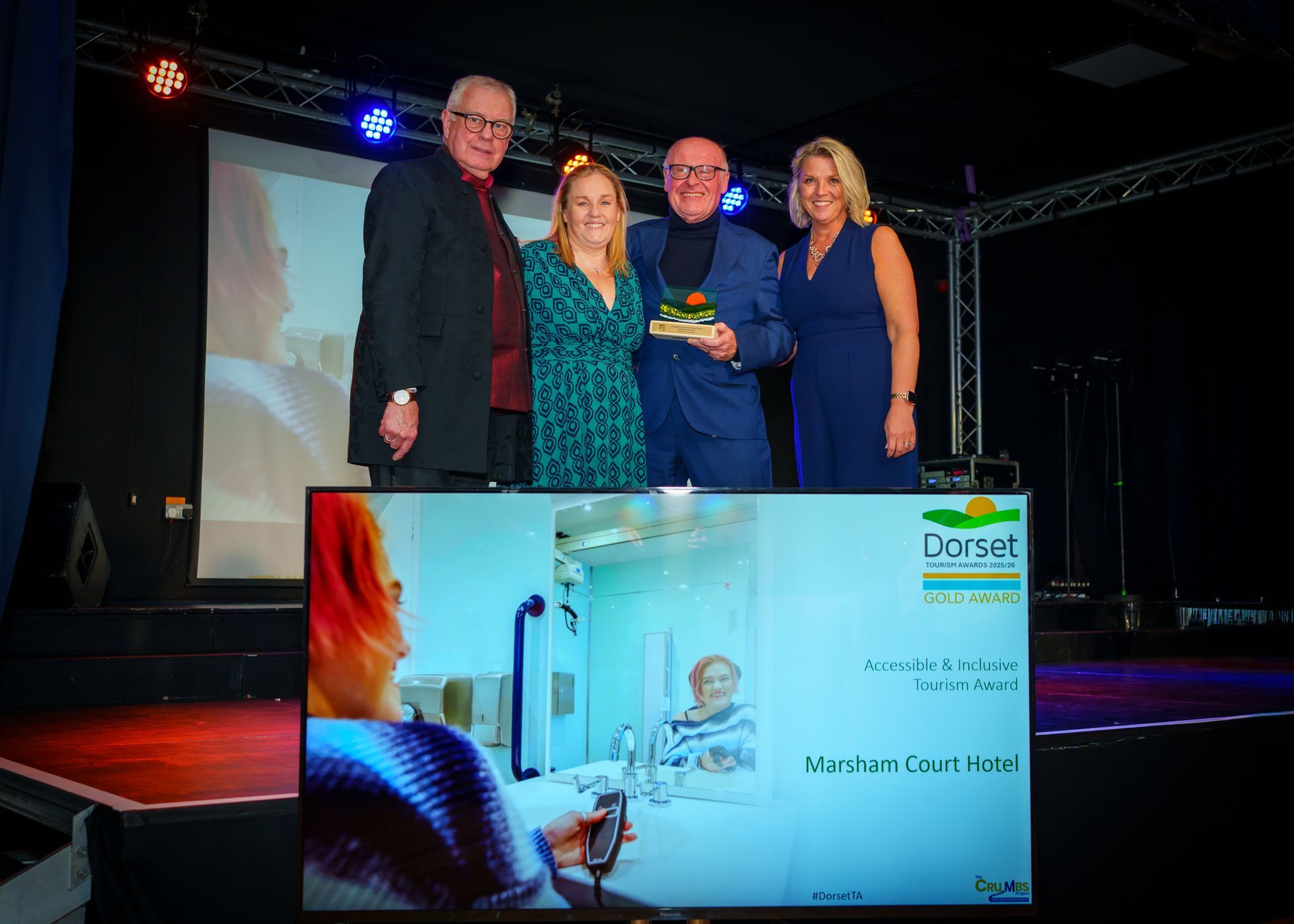Jane, Simon, Rosie and the tourist board on stage with their award with a tv screen in front which says Marsham Court Hotel Accessible & Inclusive Award. 