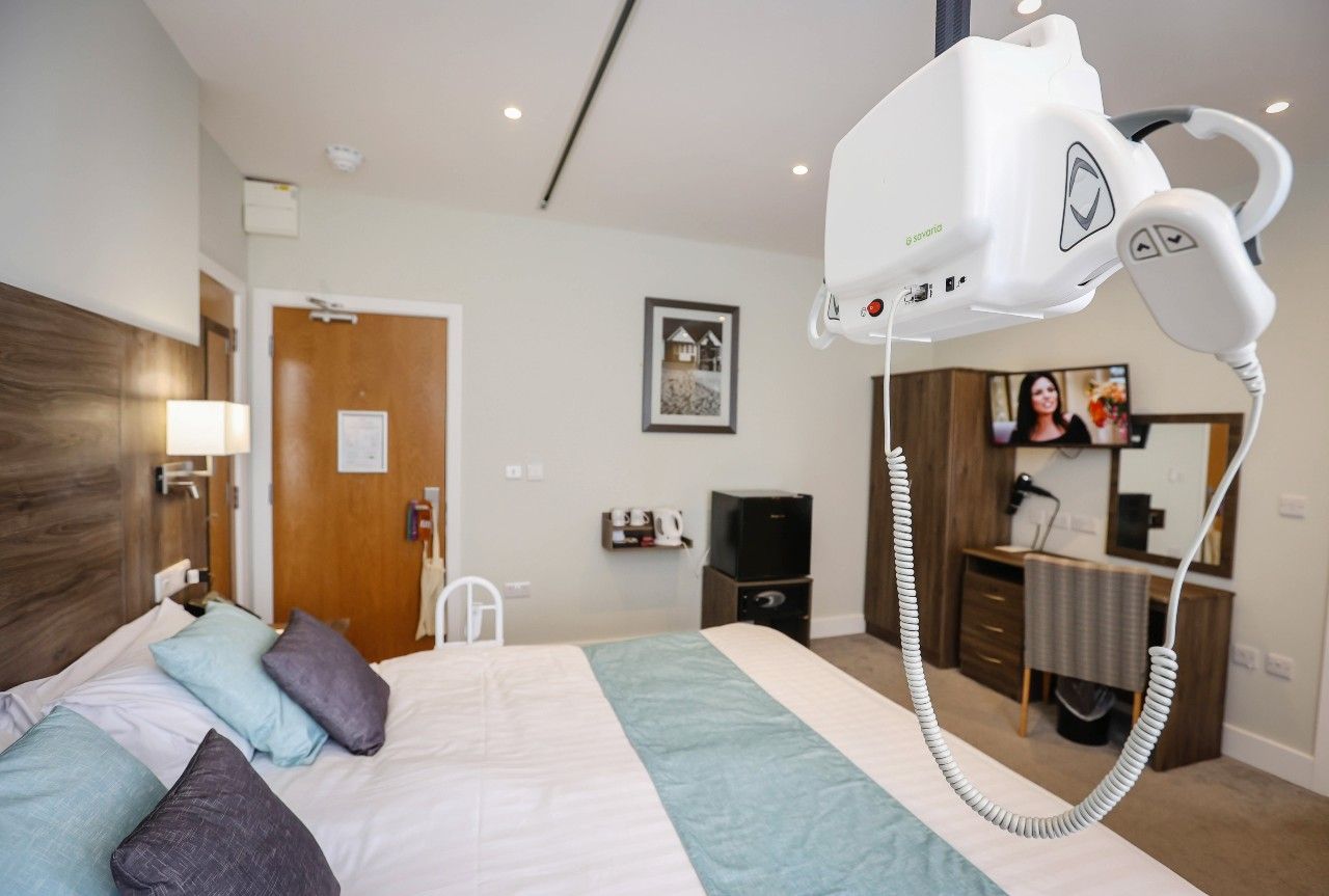 A bedroom with hoist over the bed and grab rails at the side. at the end of the bed is a wardrobe, best of draws and tv on the wall. 