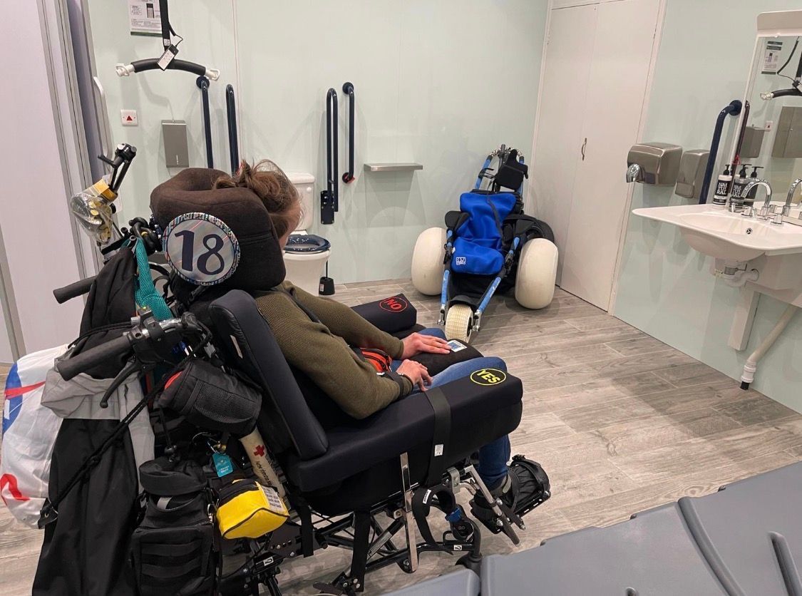 Alfie sits in his power chair looking at the changing places toilet with a beach wheelchair in the corner. 