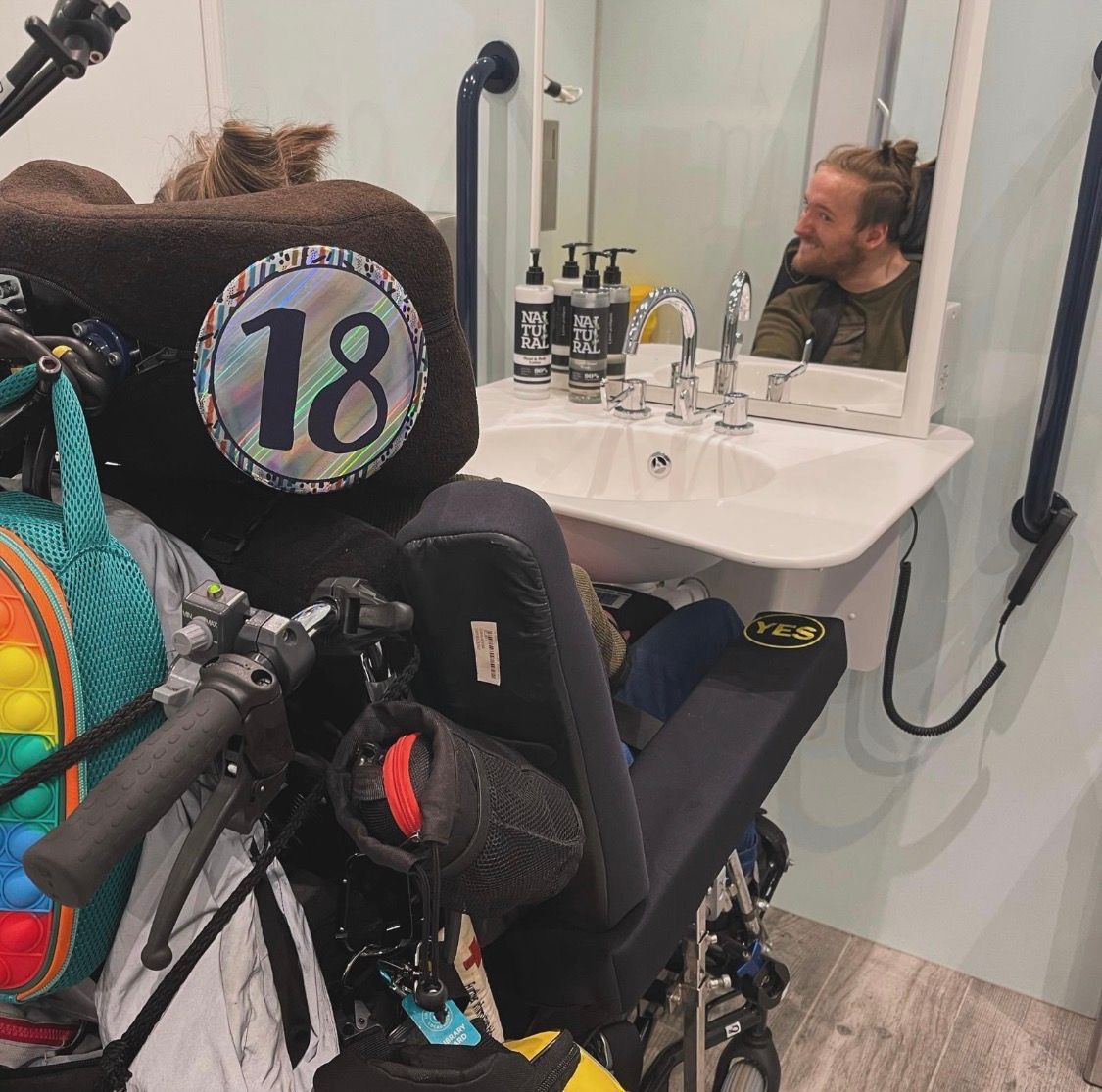 Alfie sits in his power chair with an 18 badge attached to the headrest and is looking in the mirror of the adapted bathroom of the hotel. 