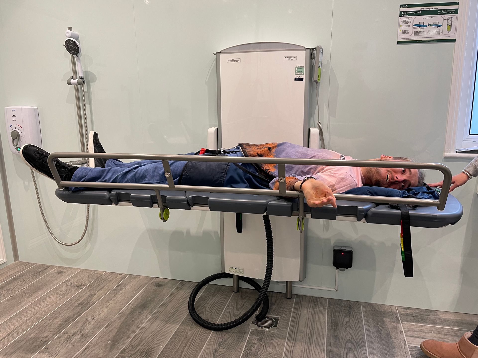 Alfie lies on a table which has remotes and a shower at the end, it is located in a change places facility. 