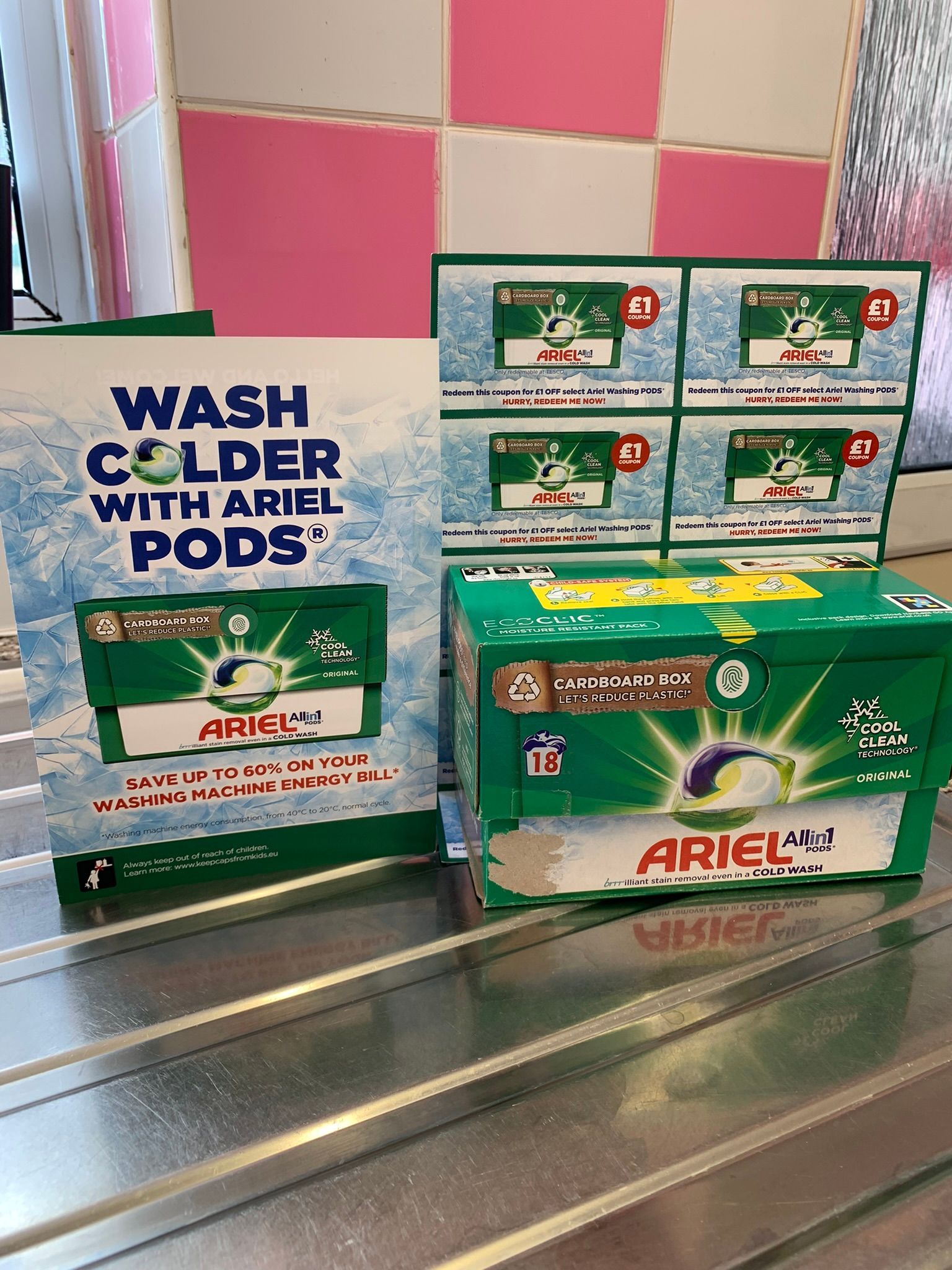 Box of ariel washing tablets in a cardboard box with a leaflet stood up next to it say wash colder with ariel pods save upto 60% on your washing machine energy bill. Behind the ariel box is some cupons for £1 off your next box. All of these products are stood on a silver draining board with pink and white tiles in the background.