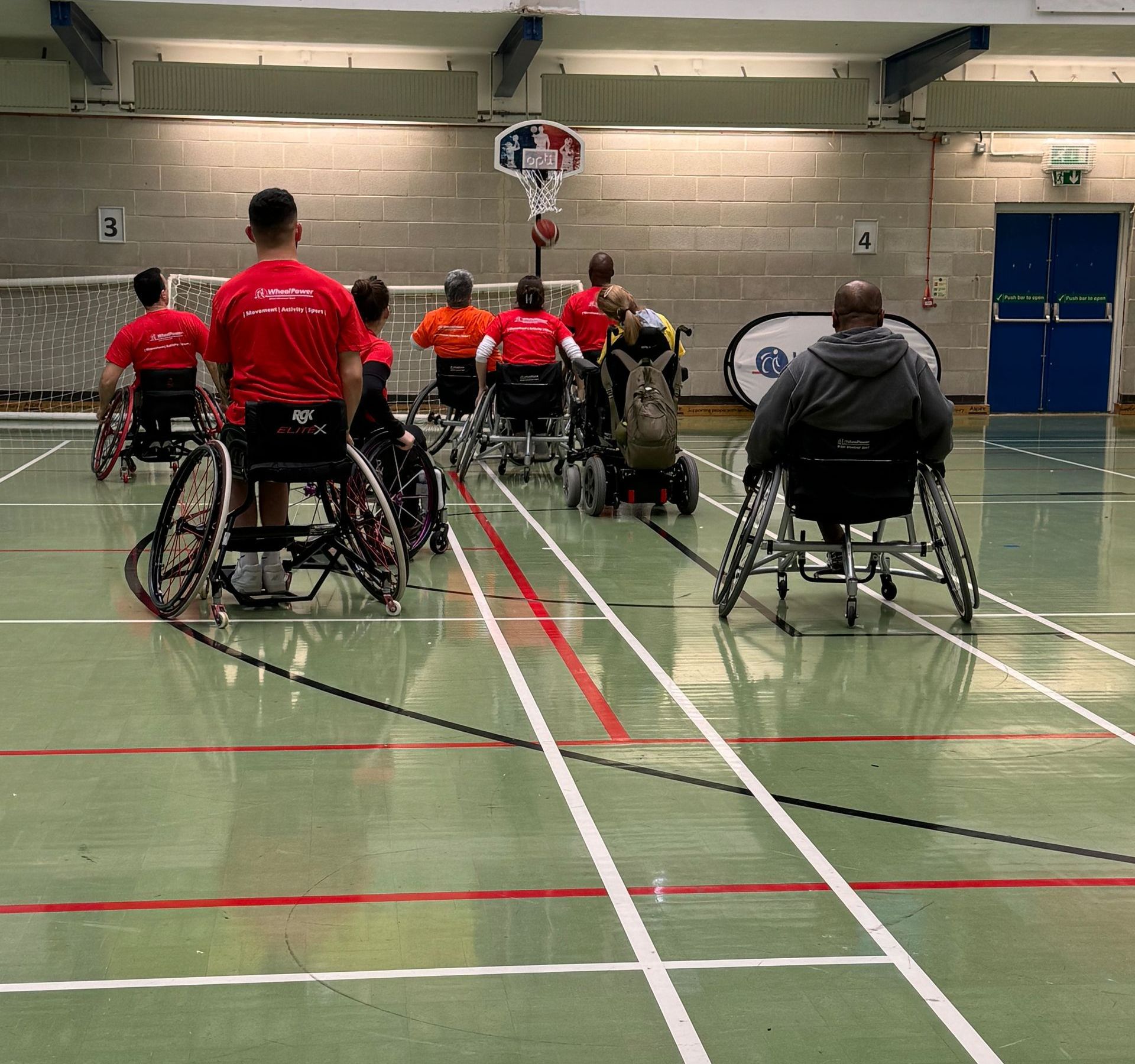 Sports Hall hosts basket ball game. Participants are wheelchair users. The net is on the back wall.