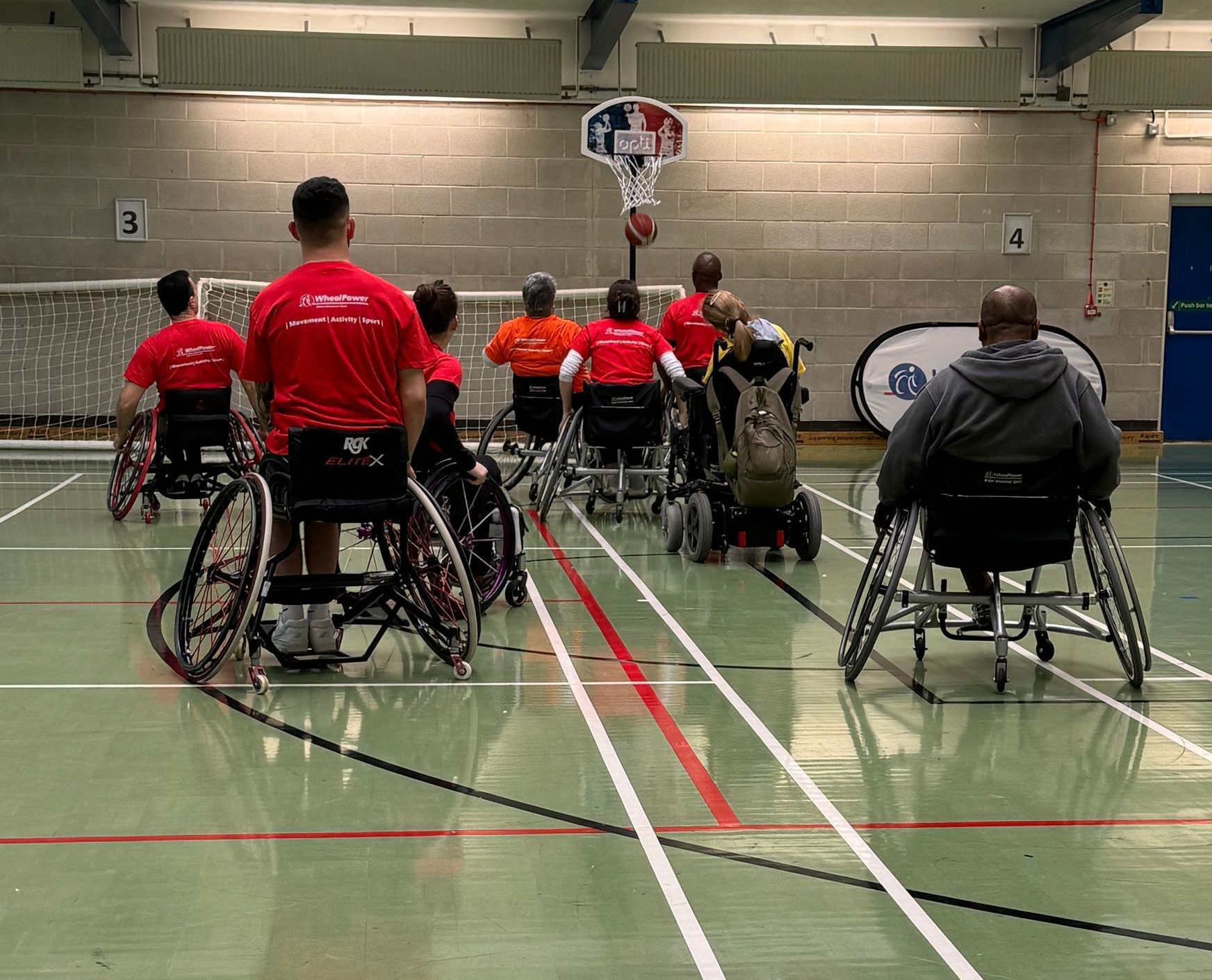 A sports hall with wheelchair users playing basketball. The net is sitting on the back wall. 