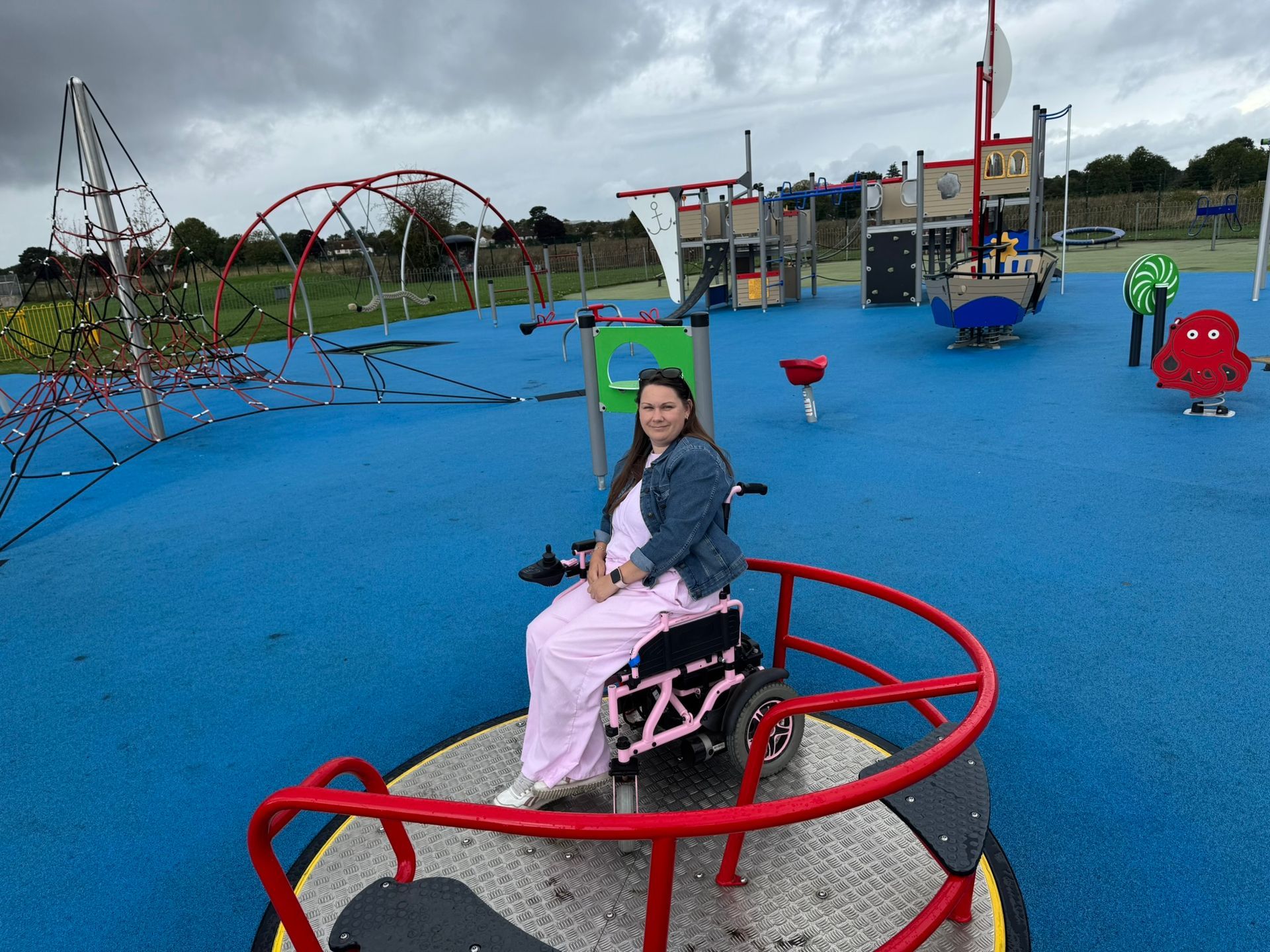 Victoria sits in here wheelchair on a red wheelchair accessible roundabout in a children's playground. 