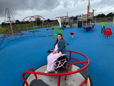 Victoria sits on a red wheelchair accessible roundabout in a children's playground with a blue floor. 