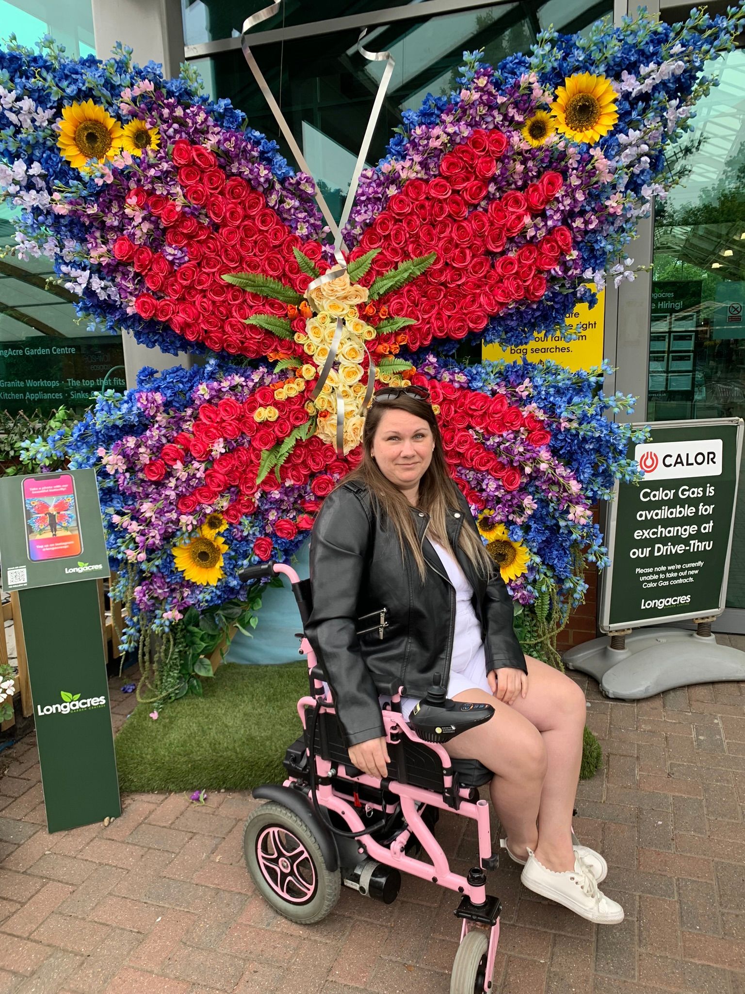 White Female with brown hair - wearing a black leather jacket and  is a powerchair user which is a pink colour.