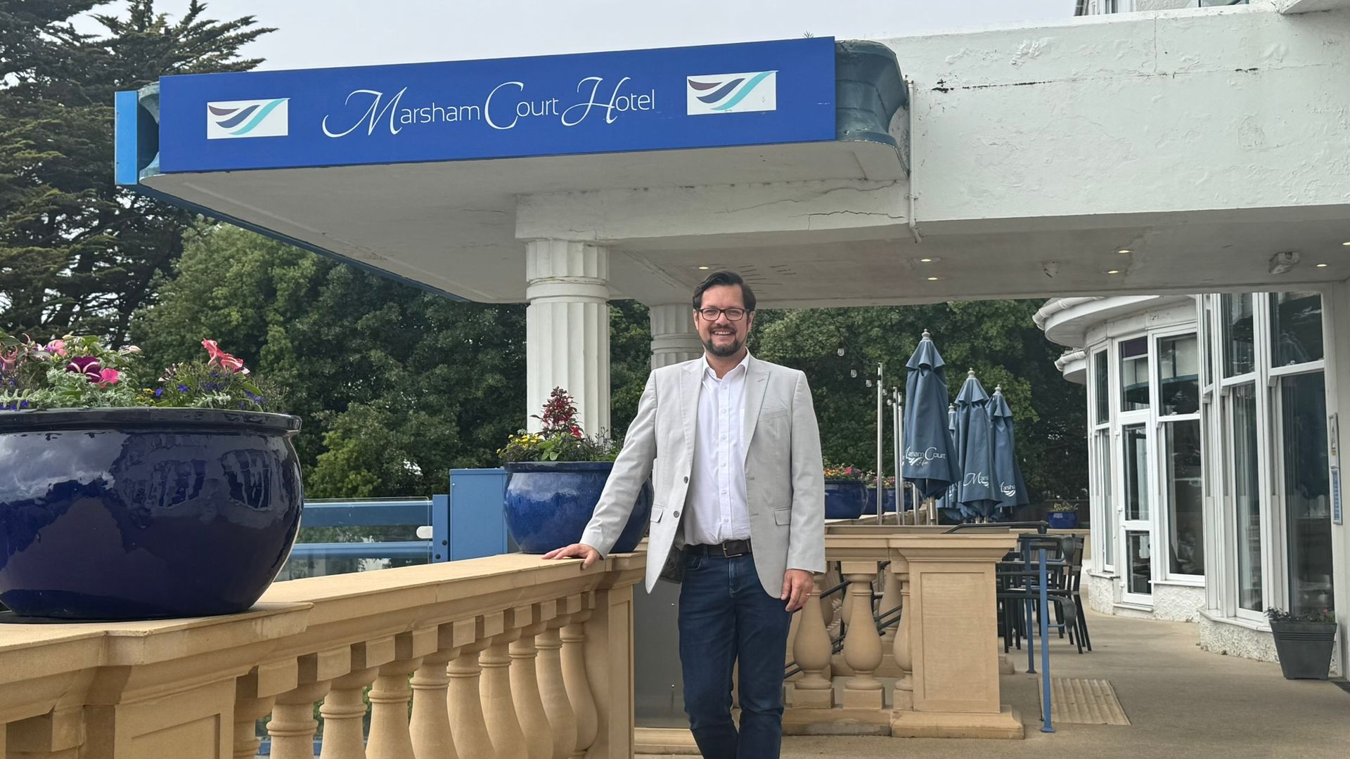 James stands with his hand on the beige wall of the terrace of the hotel. Blue flower pots with pink