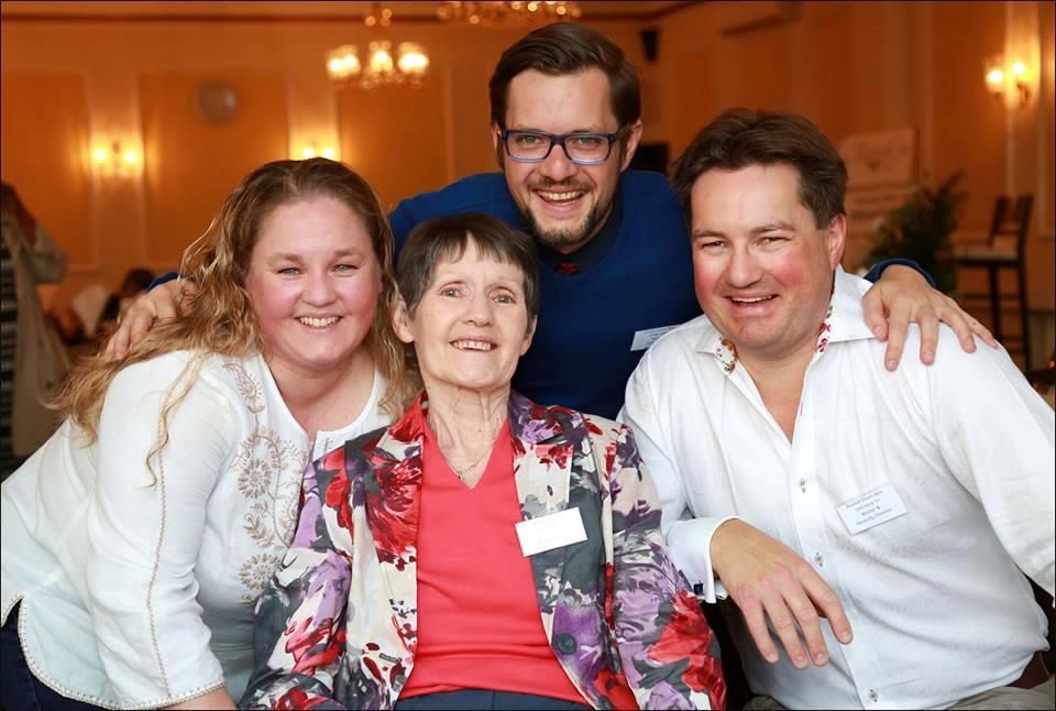 Jeannie Deavin sits in the middle with a red blouse and multicoloured jacket on her children surround her leaning in with smiles on their faces. 