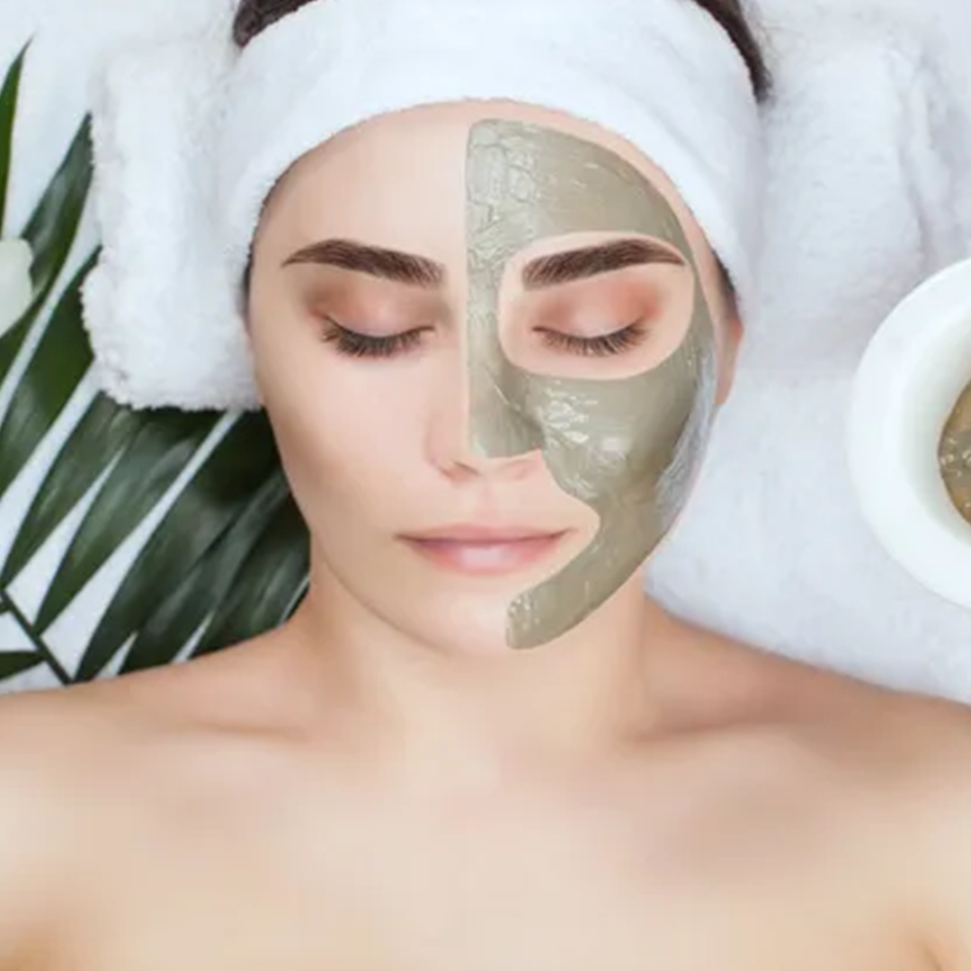 Woman with half her face covered in a mud mask, relaxing with a towel on her head.